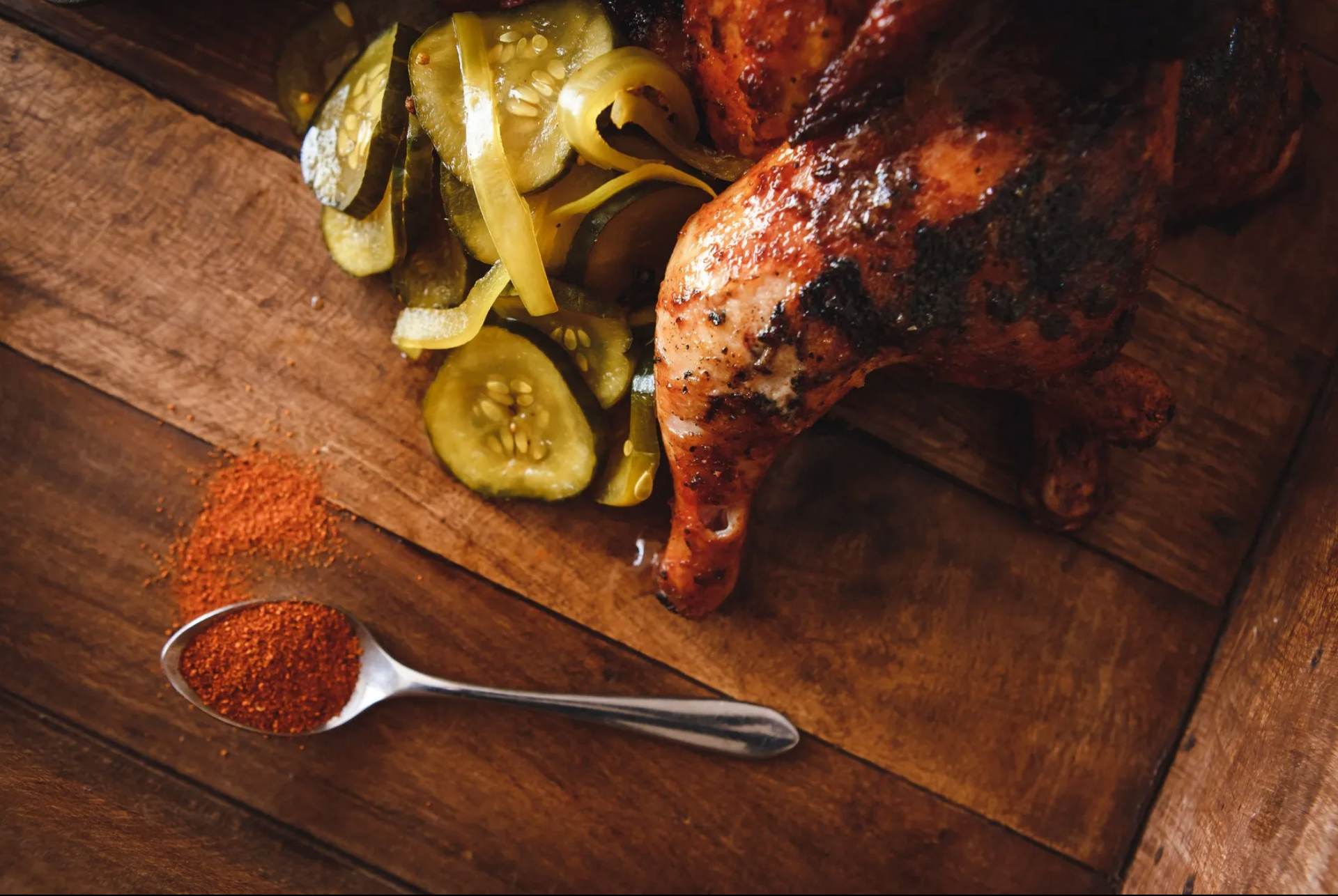 A wooden table topped with chicken , pickles and a spoon of seasoning.