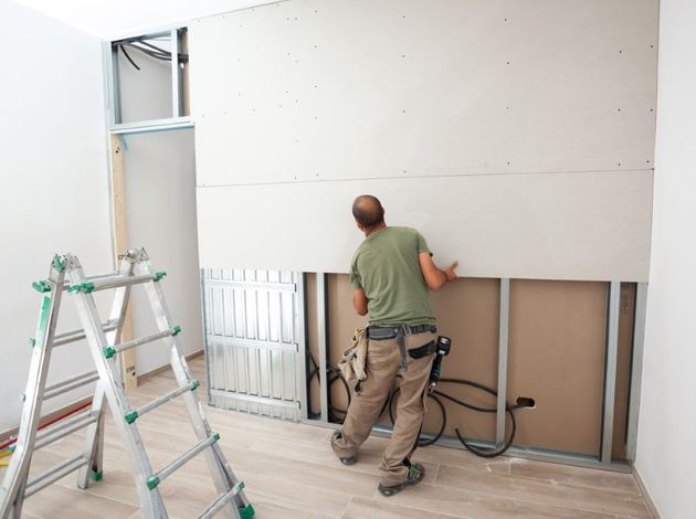 Worker installing drywall in a room, with a ladder and exposed framing visible.