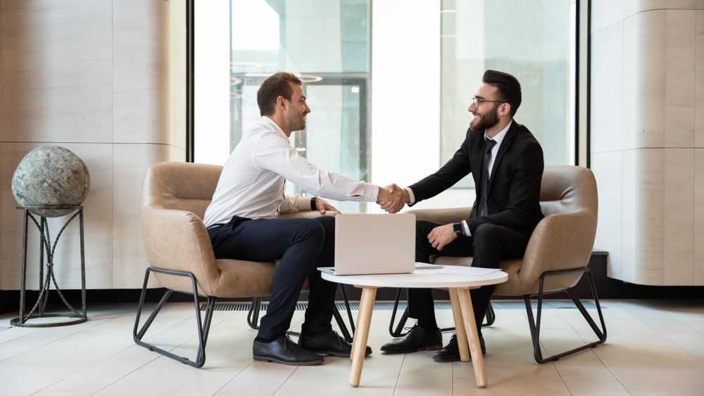 Two men in business attire shaking hands, smiling, seated in a modern office with a laptop on a small table GMS Legal & Conveyancing In 38 Canton Beach Rd, Toukley
