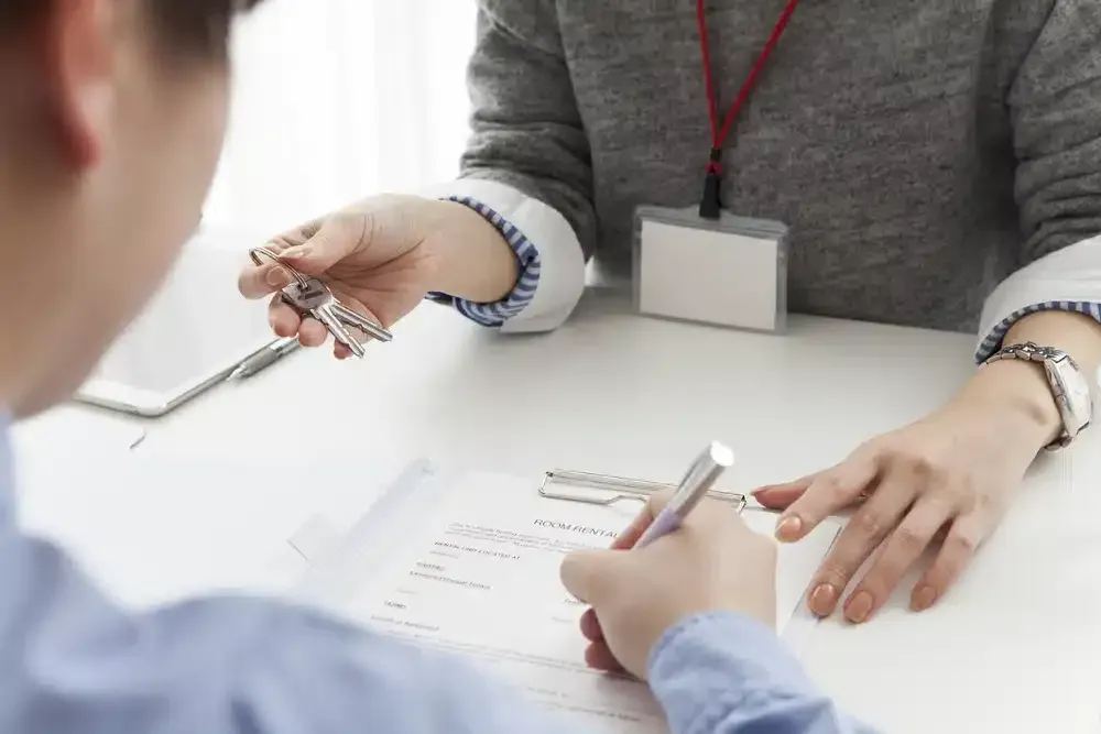 Person signing a document while another hands over keys at a desk. GMS Legal & Conveyancing In 38 Canton Beach Rd, Toukley
