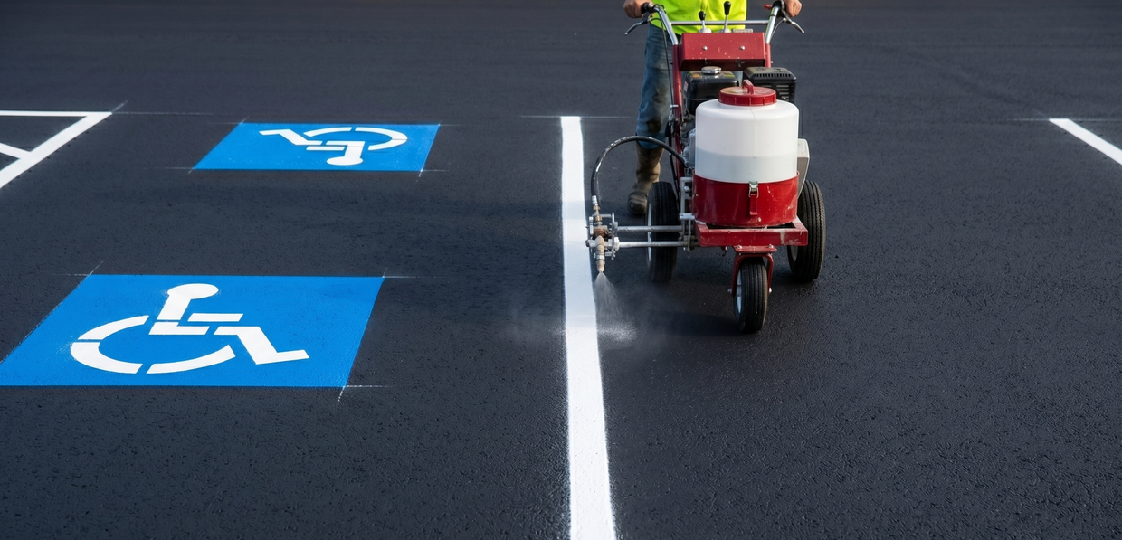 A worker marking white handicap parking symbols on a dark asphalt road with a line-painting machine.