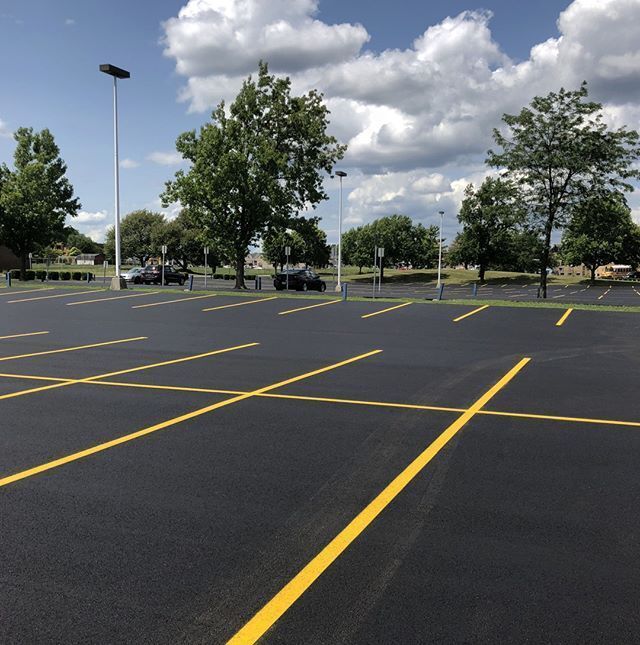 Empty parking lot with yellow lines under a partly cloudy sky, trees and light poles in the background