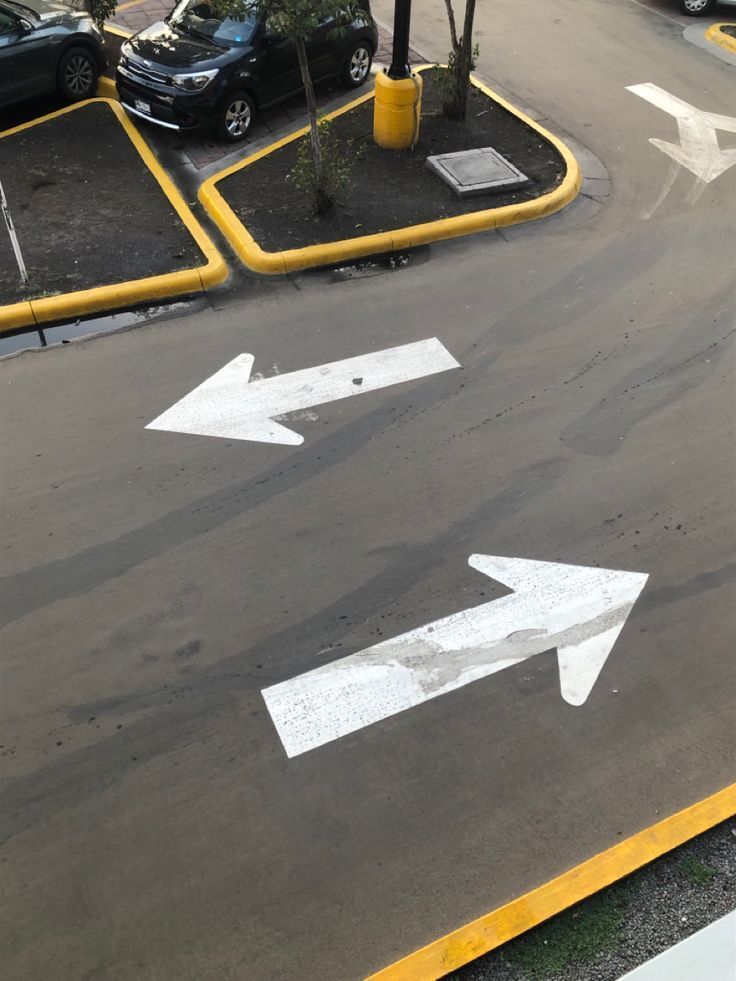 Parking lot intersection with two large white directional arrows painted on dark pavement, edged in yellow.
