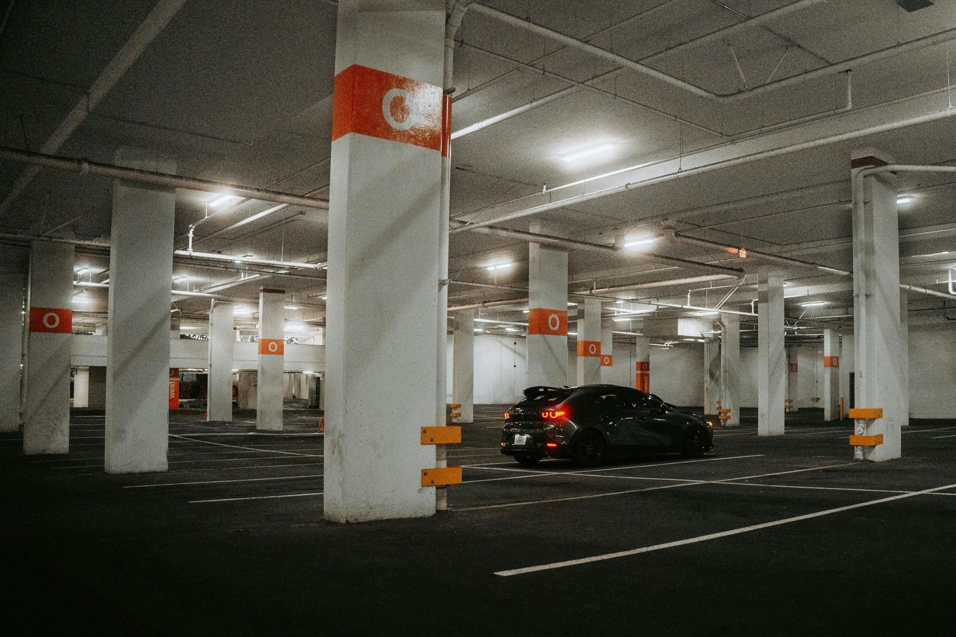 Dimly lit parking garage with a black car and white columns marked by orange signs