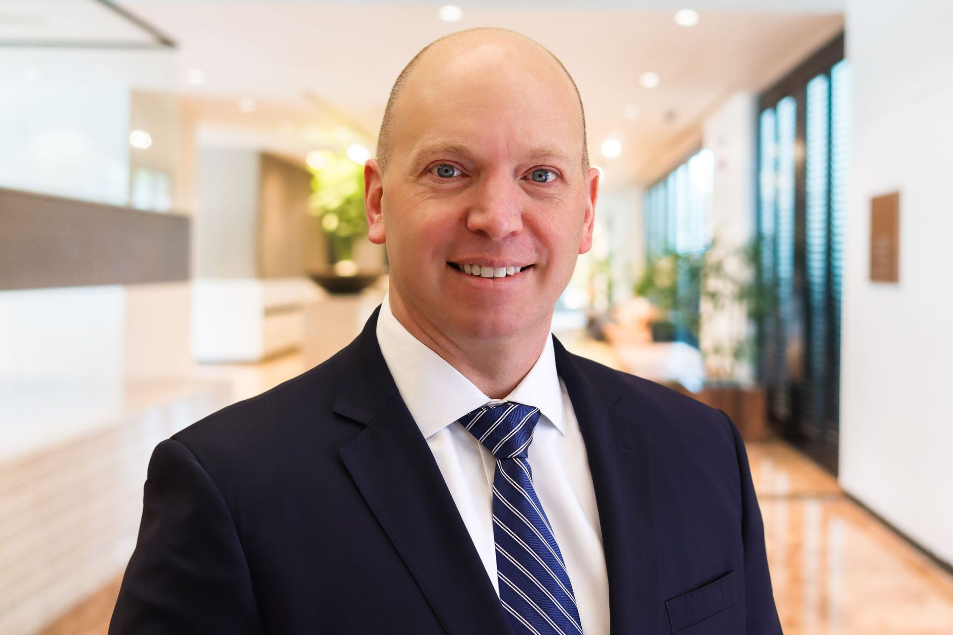 Smiling bald man in a navy suit and striped tie standing in a bright office hallway