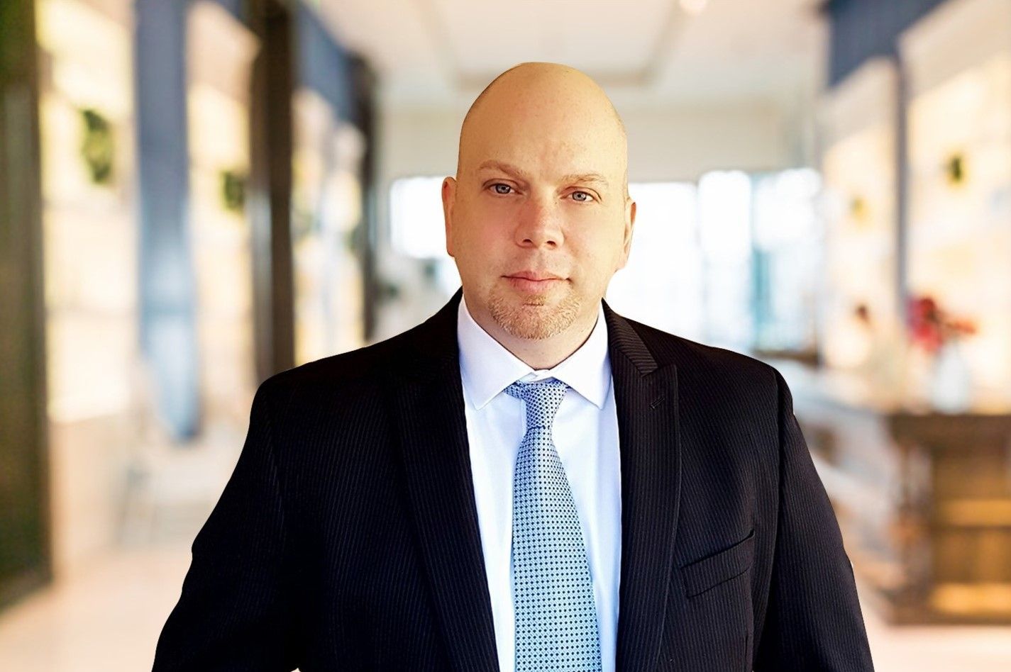 Portrait of a man in a dark suit and blue tie standing in a bright hallway