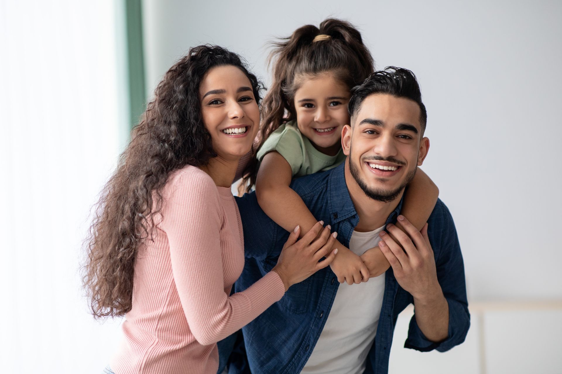 Smiling family of three posing indoors against a light background