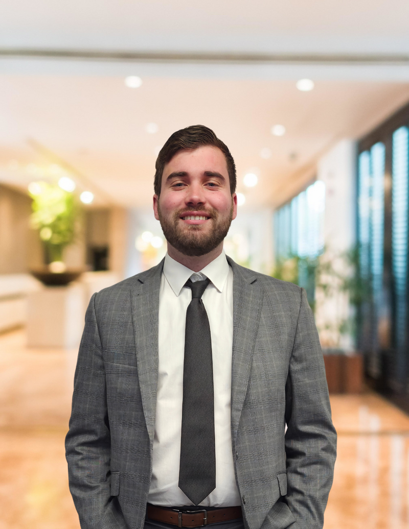 Man in a gray suit and tie standing in a bright office hallway, smiling at the camera.