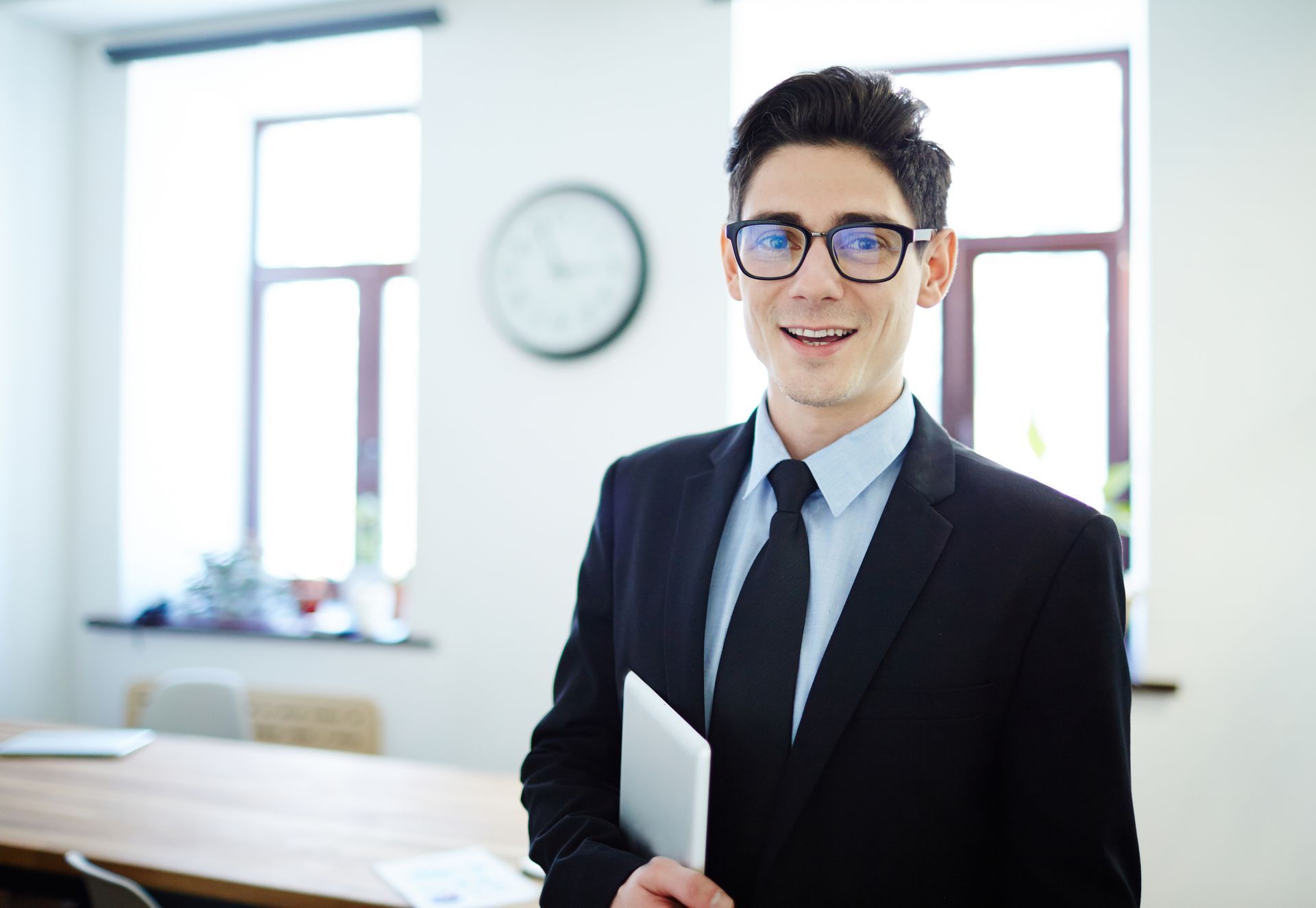 Smiling professional in a black suit holding a folder in a bright office by a window and wall clock