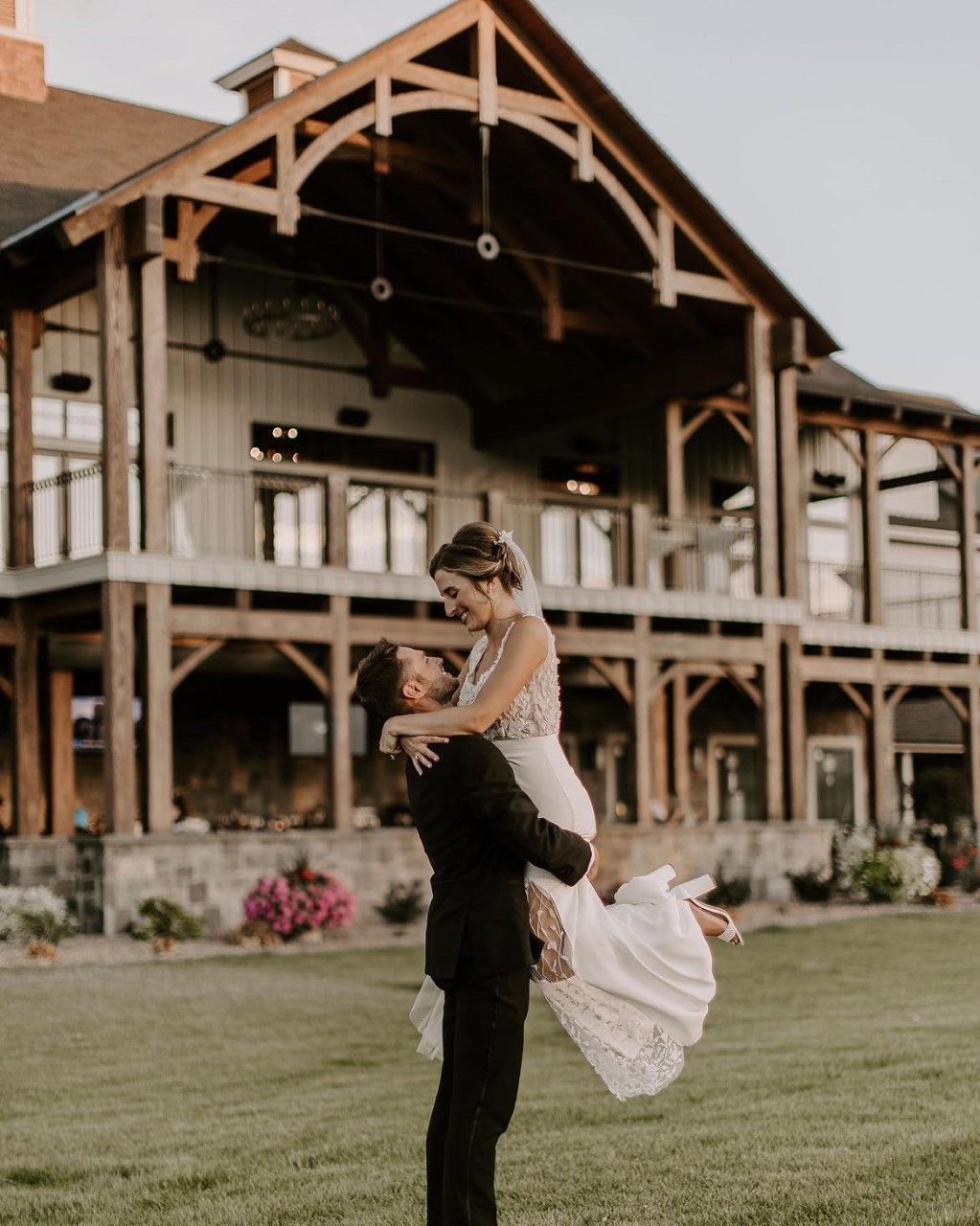 Groom lifts bride in front of a large wooden building; both smiling; sunny outdoor setting.