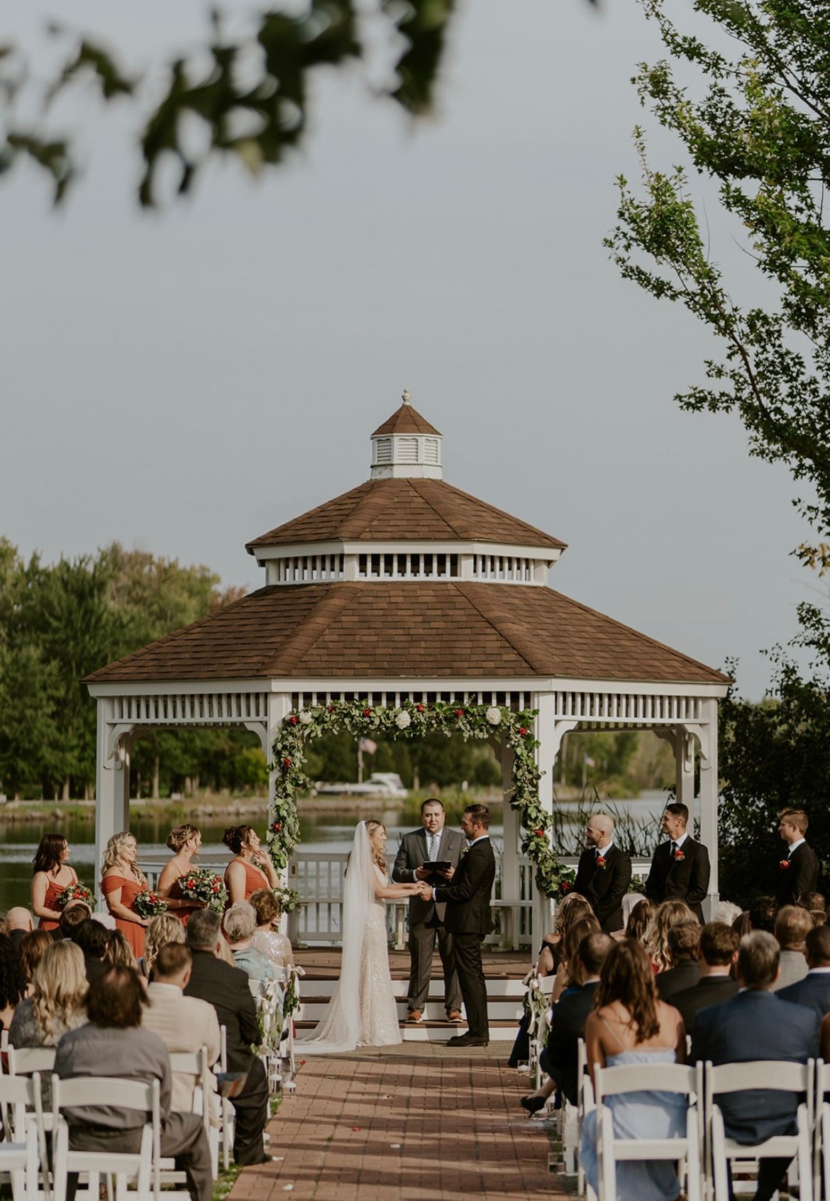 Wedding ceremony near a gazebo by a lake. Bride and groom hold hands, guests watch.