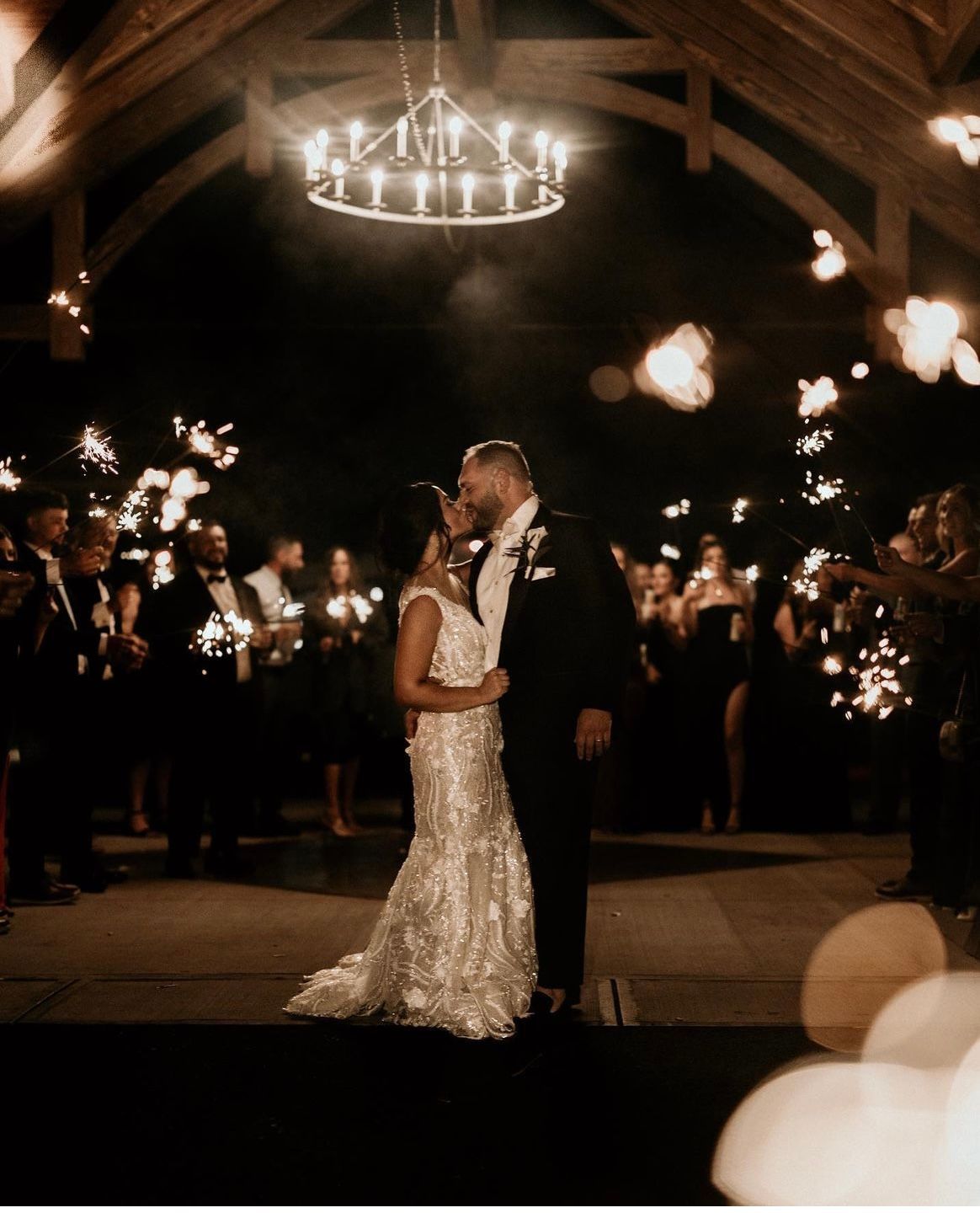 Bride and groom kiss amidst sparklers held by wedding guests, lit at night under a chandelier.