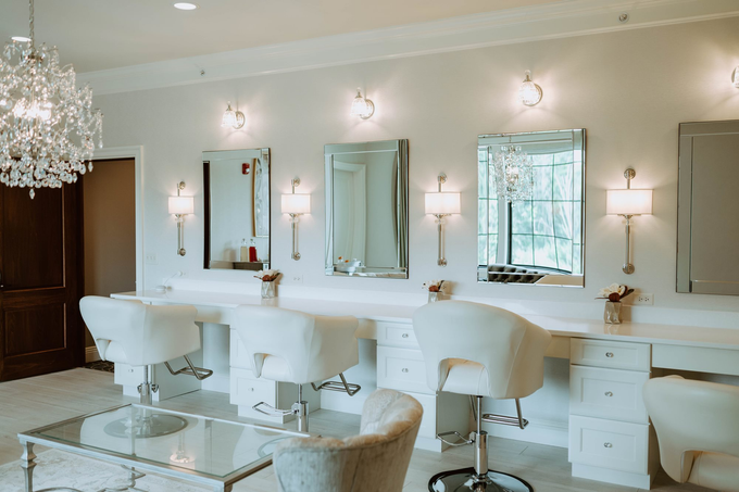 Hair salon with white vanities, mirrors, and chairs, with a crystal chandelier and wall sconces.