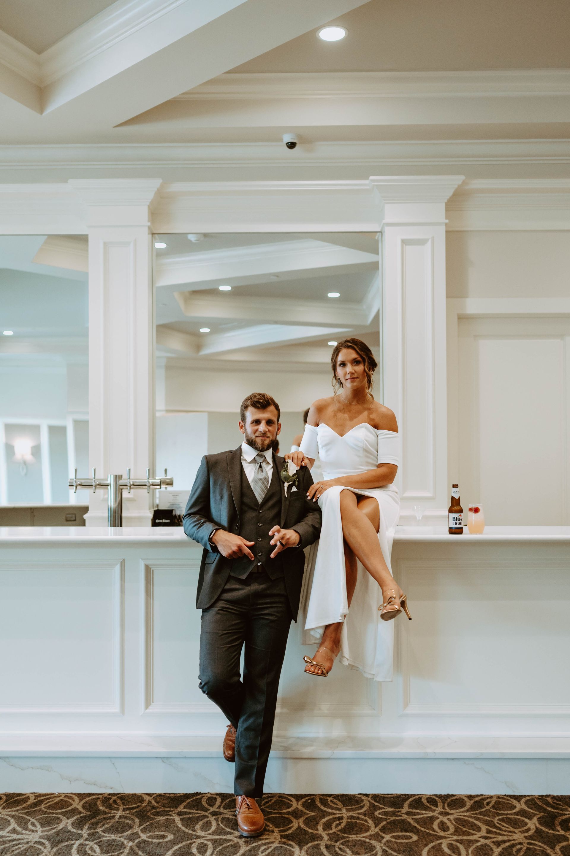 Newlyweds pose at a bar. Woman in white dress sits, man in suit stands, both smile. Modern, white interior.
