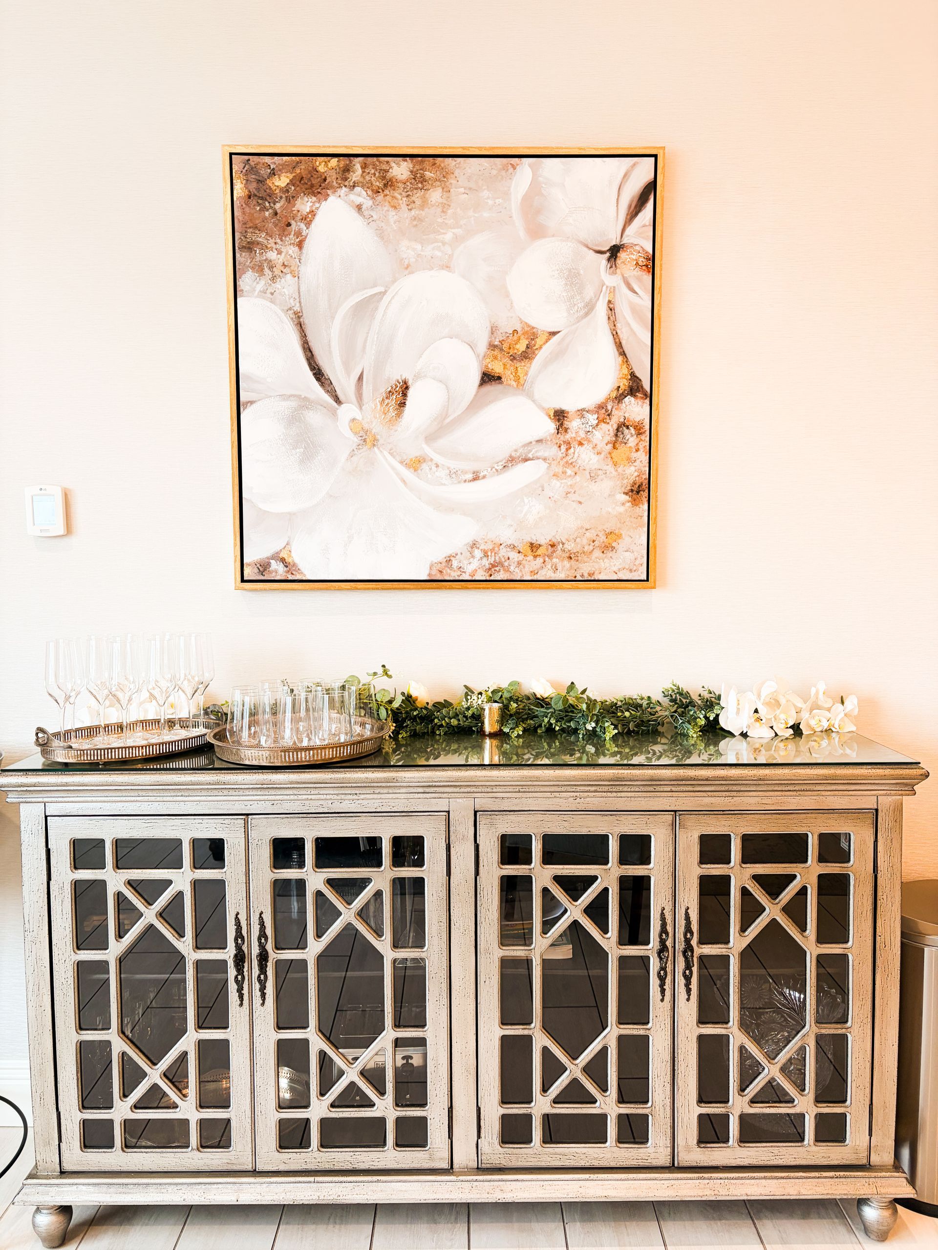 White cabinet with glass-paneled doors, topped with decor and a floral print painting above.