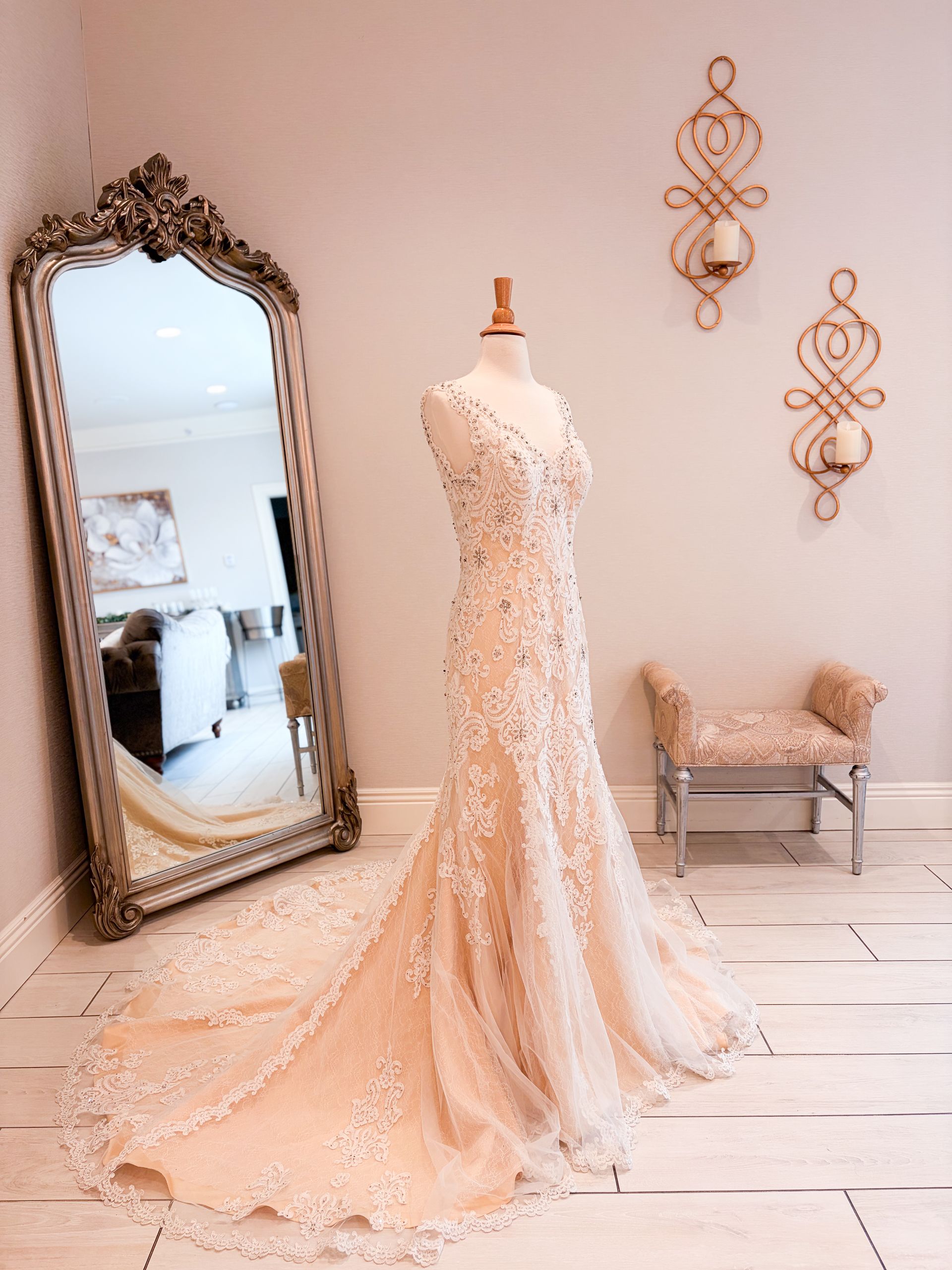 Wedding dress on a mannequin in a light room with a mirror and decorative wall sconces.