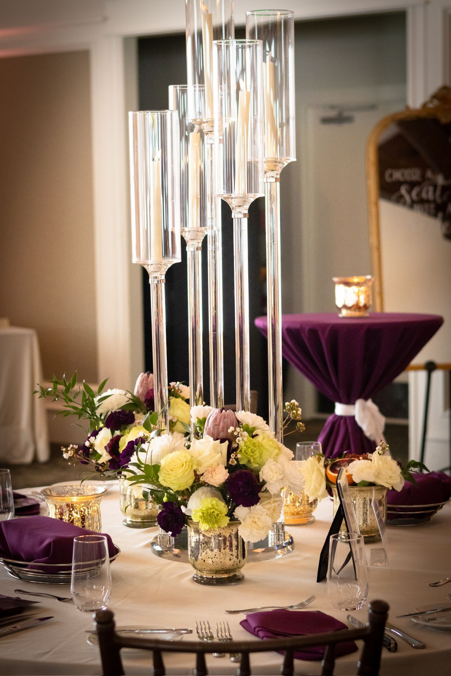 Table setting with floral centerpieces, tall candleholders, and a small purple table in the background.