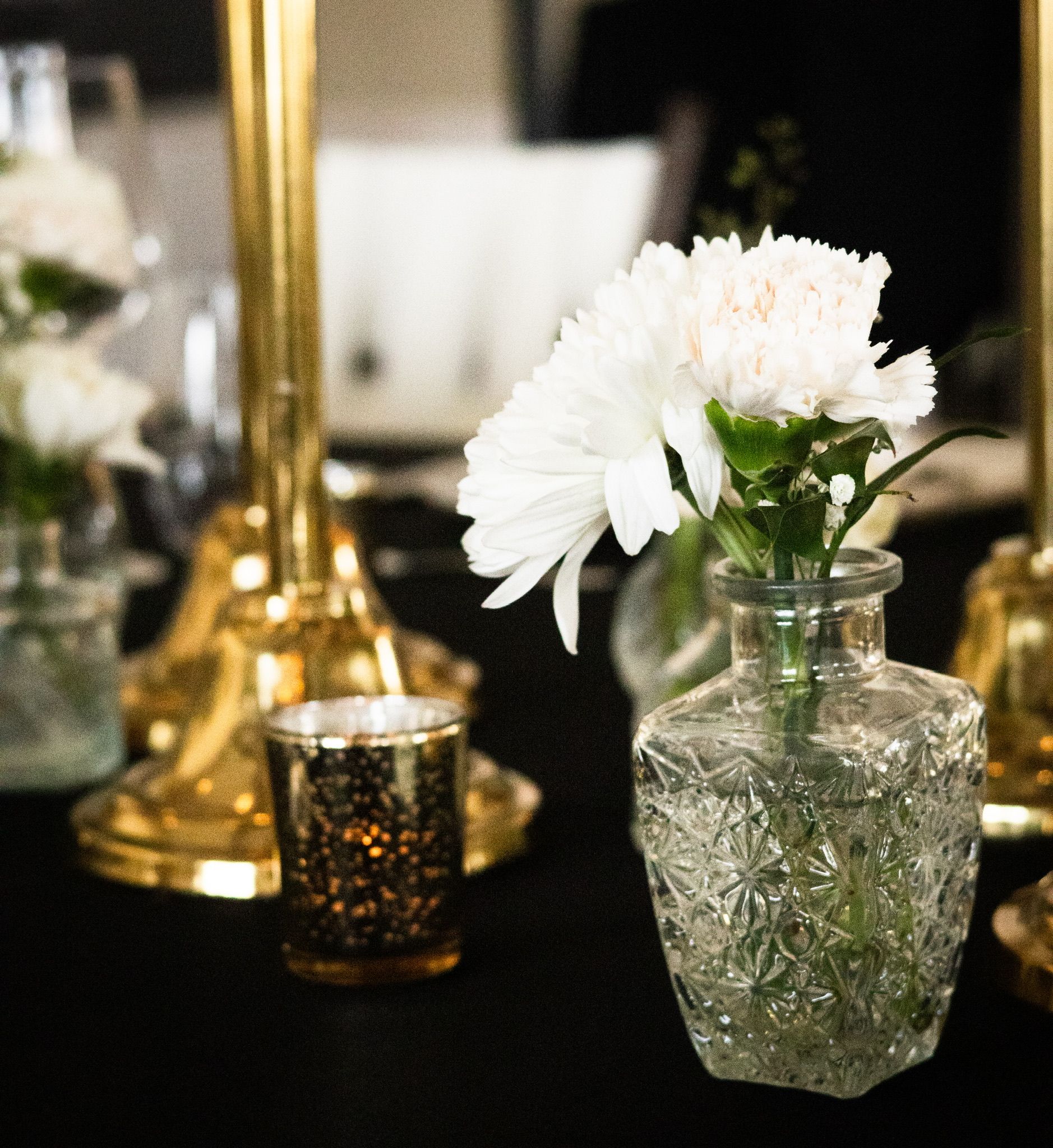 White flowers in a patterned glass vase on a table with gold accents and candlelight.