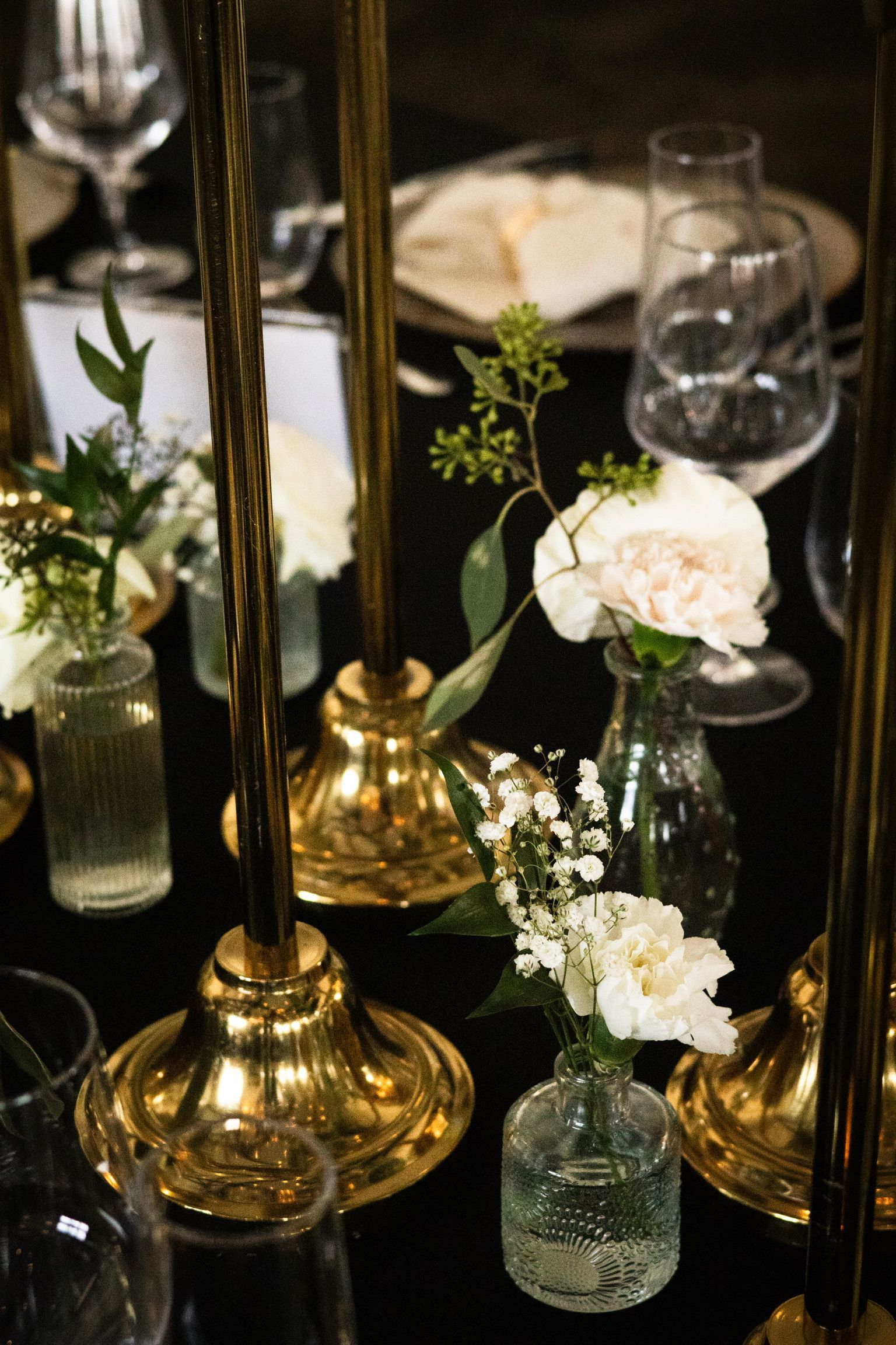 Golden candle holders and small vases of white flowers on a black table setting.