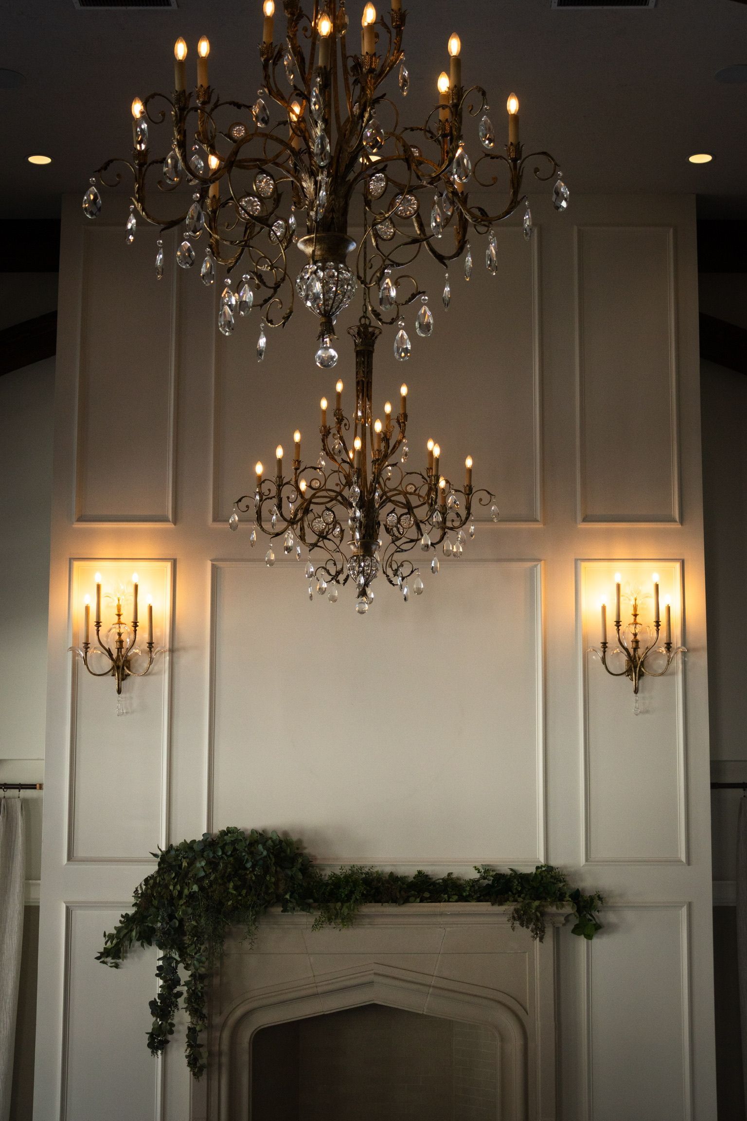 Two ornate chandeliers hang above a white fireplace with green garland, sconces on either side.
