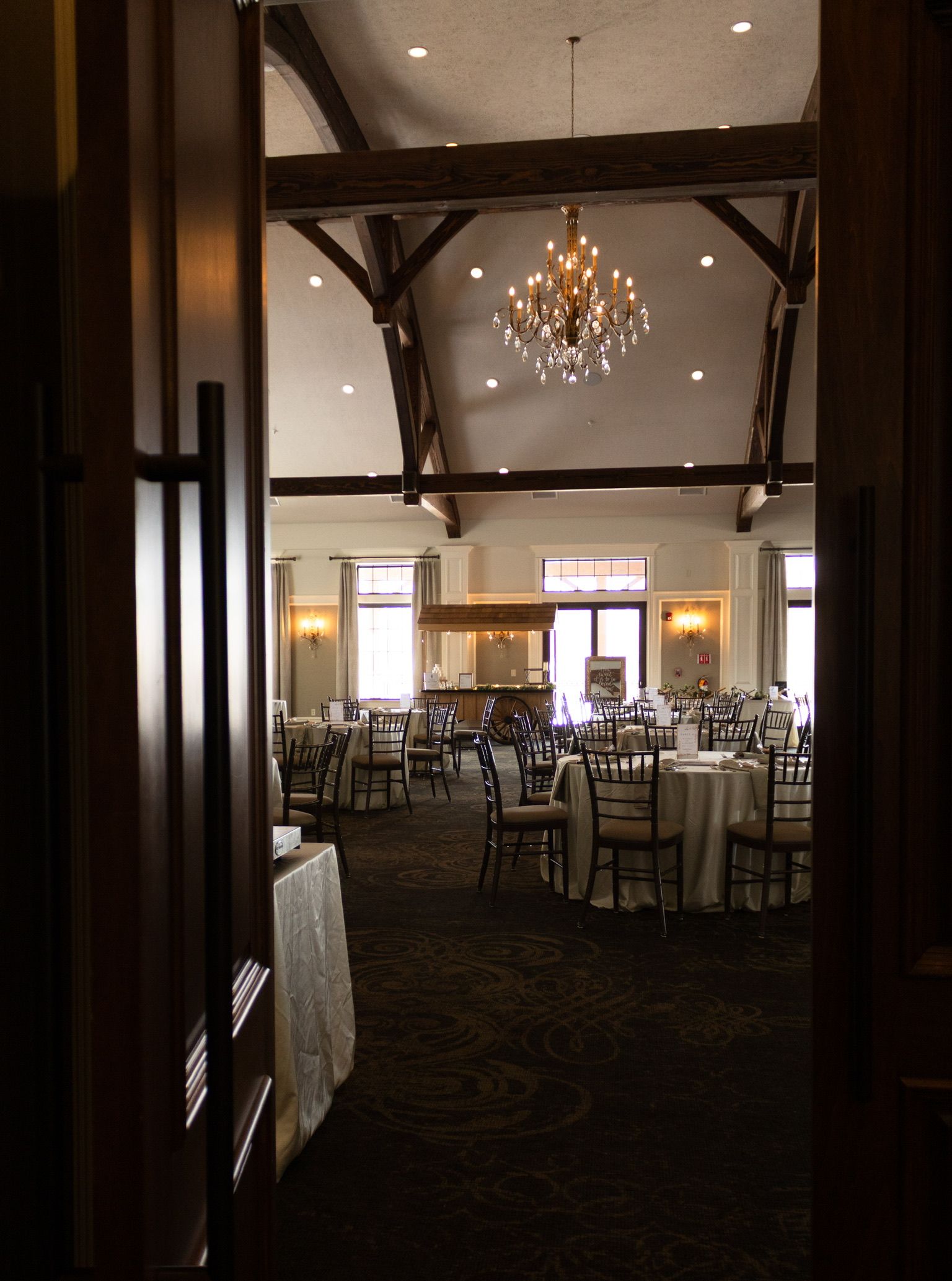 View through a doorway of a large room with round tables, a chandelier, and exposed wooden beams.