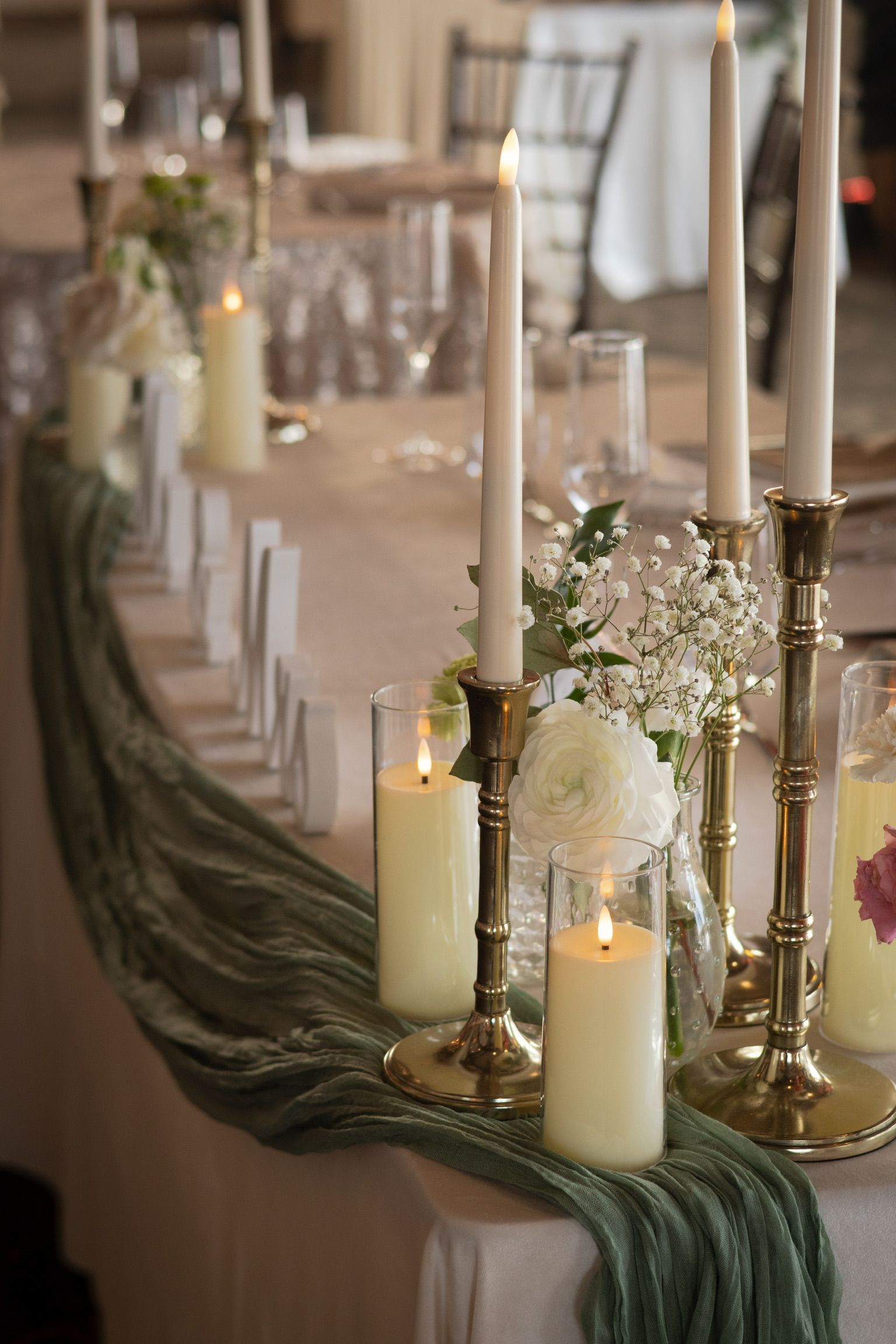 Elegant table setting with candles, greenery, and glassware, draped with a green runner, in a well-lit room.