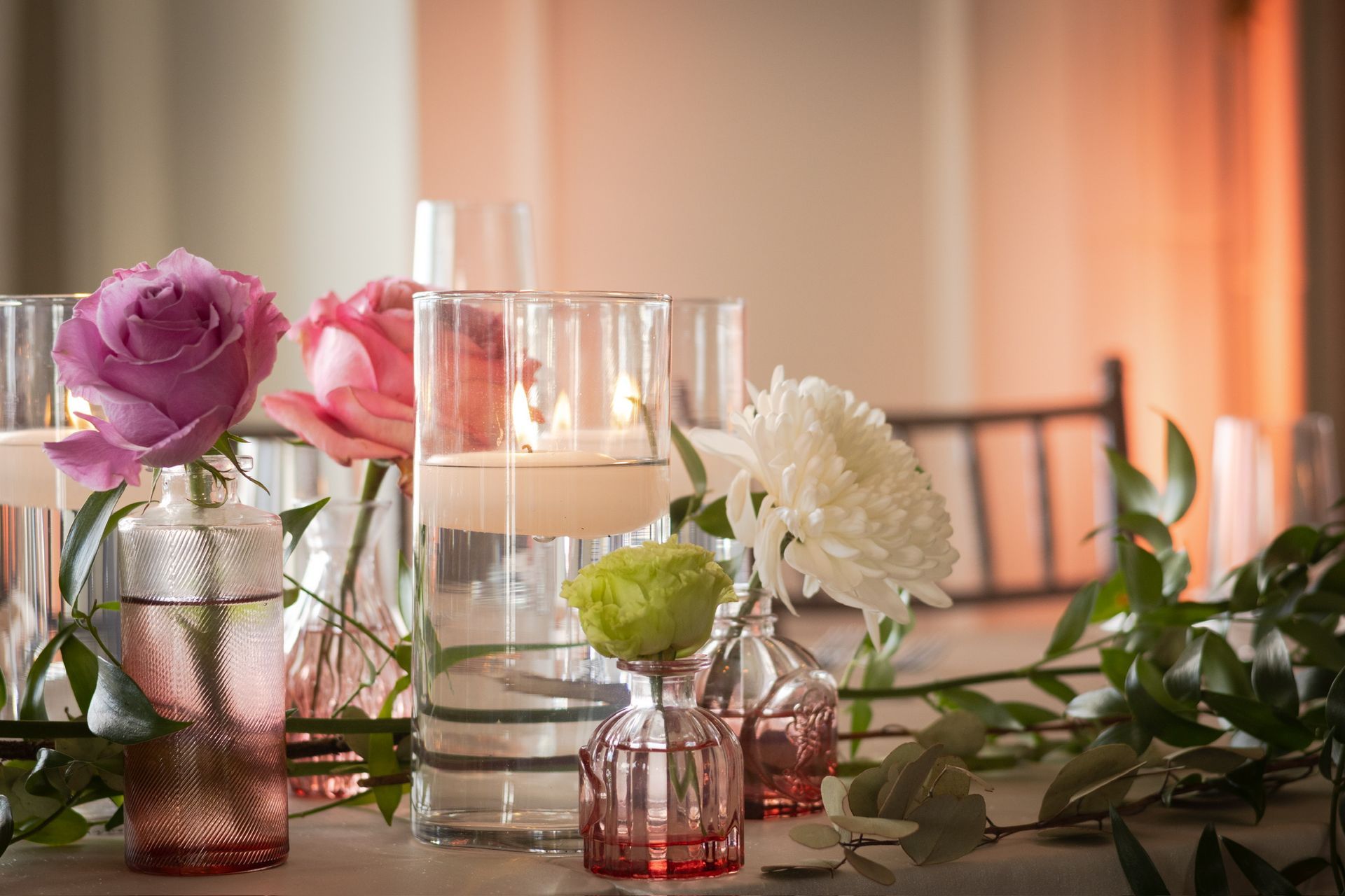 Floral centerpiece on a table: roses and chrysanthemums in glass vases, with candles and greenery.