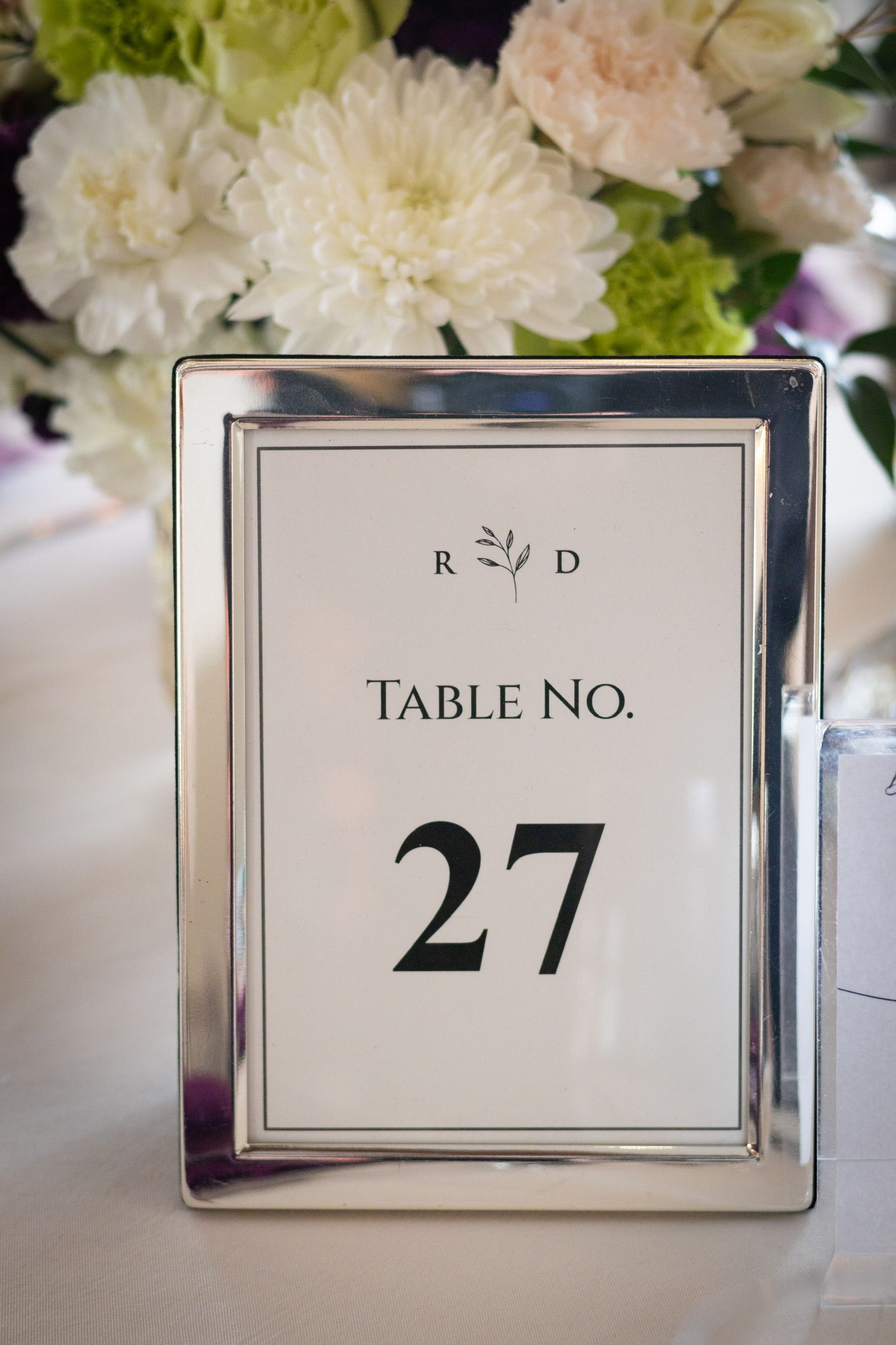 Table number 27 in a silver frame with white background, floral arrangement in the background.