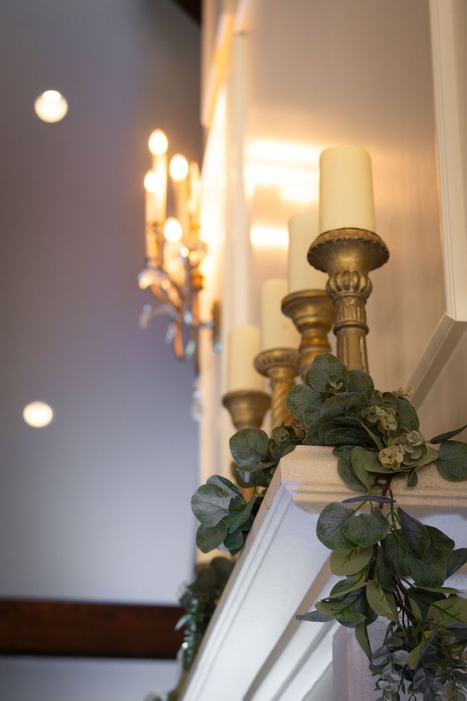 Mantelpiece decorated with gold candle holders, greenery, and lit candles, with a chandelier in the background.