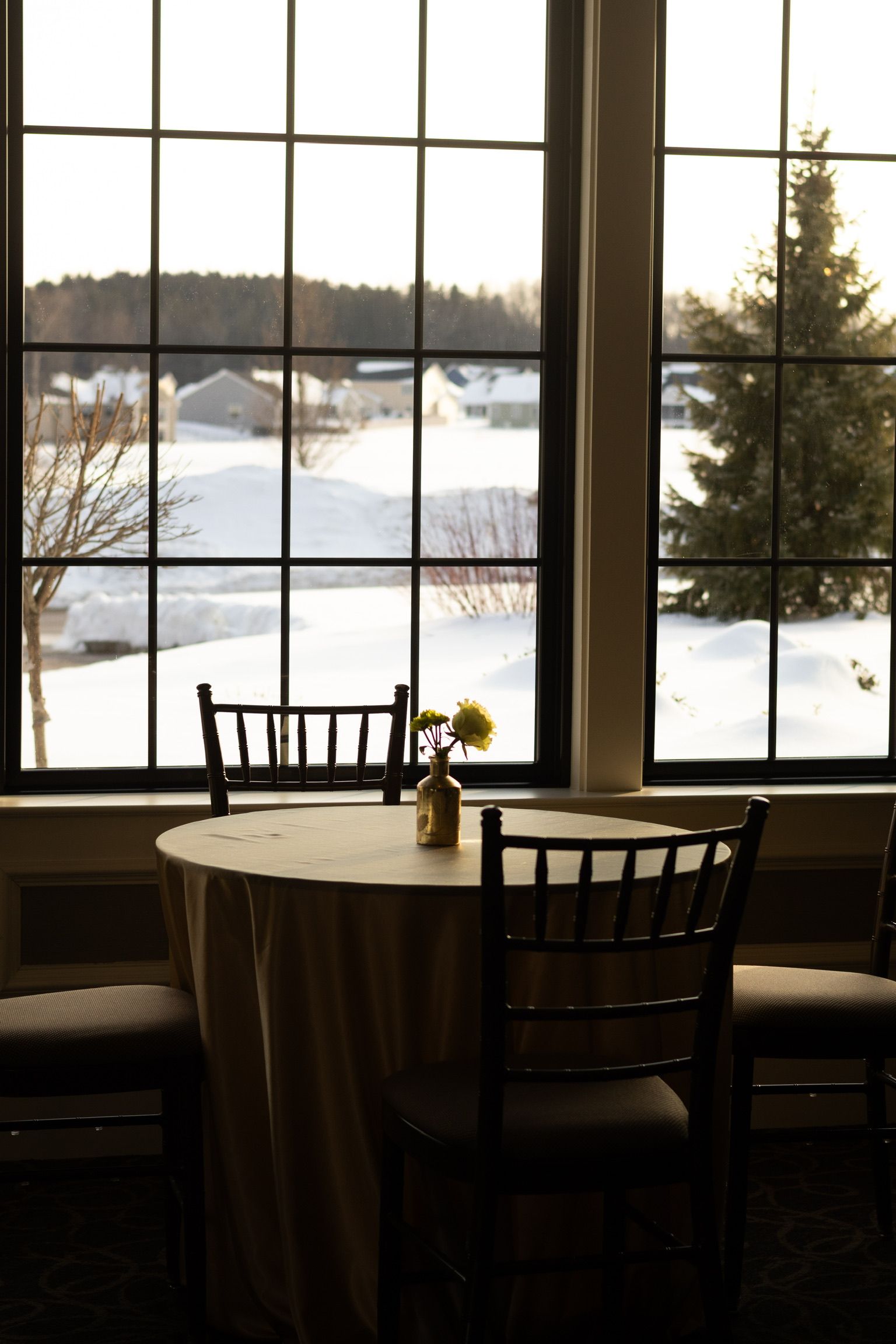 A table for two with chairs sits near a window overlooking a snowy landscape. A vase of yellow flowers is on the table.
