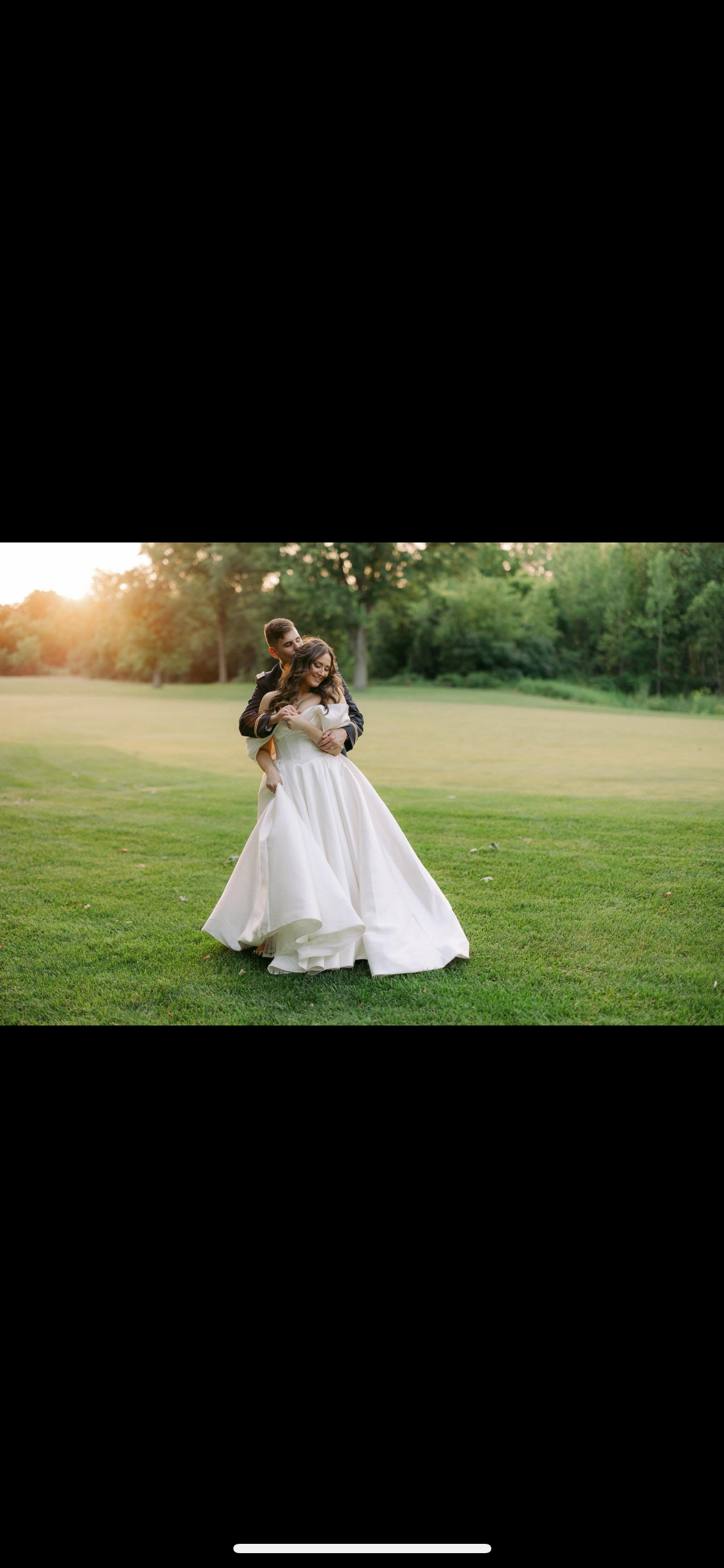 Couple embracing on a grassy field; bride in a white gown. Warm sunlight shines in the background.