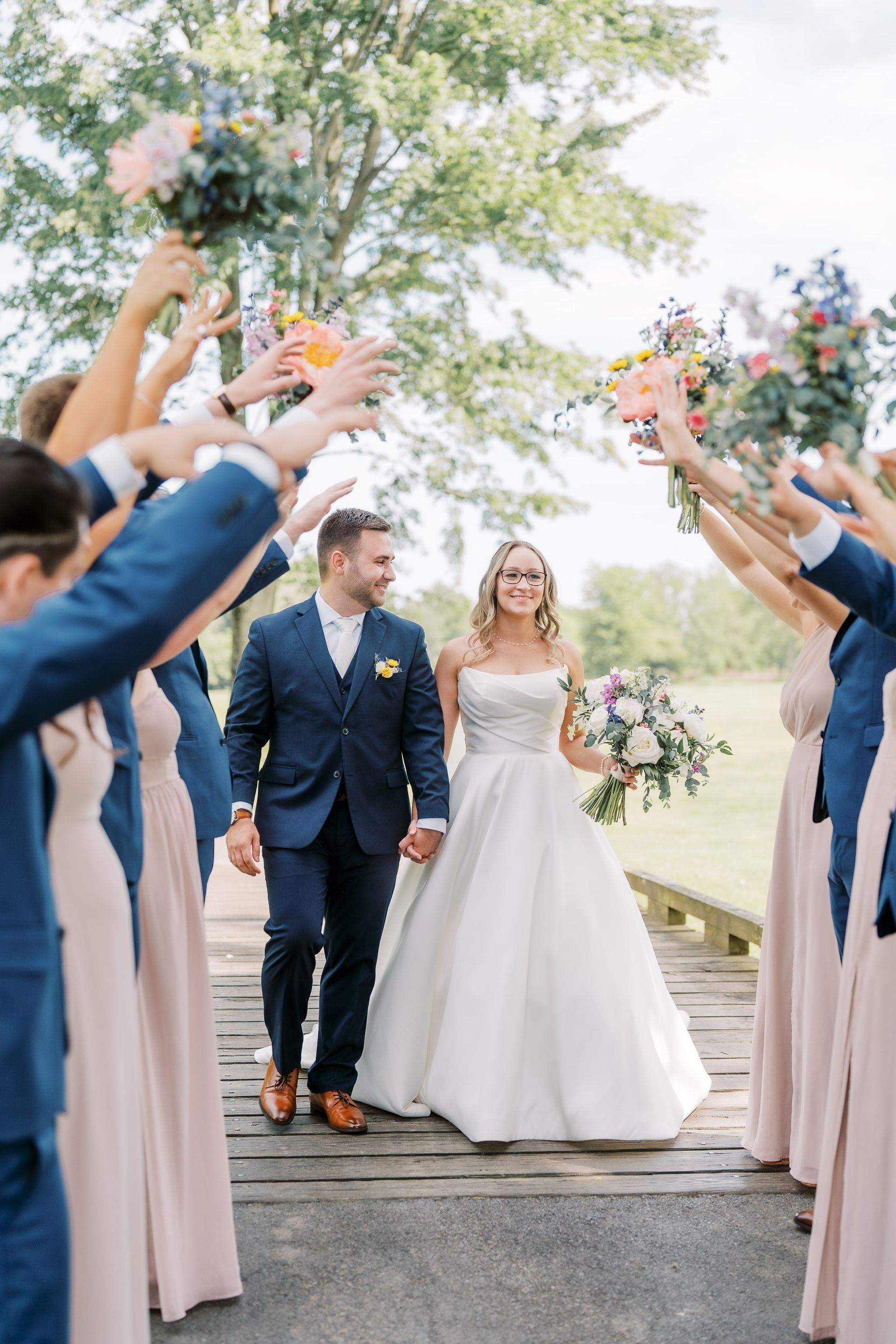Newlyweds walk hand-in-hand through an arch of raised arms, smiling. Wedding party cheers, outdoors.