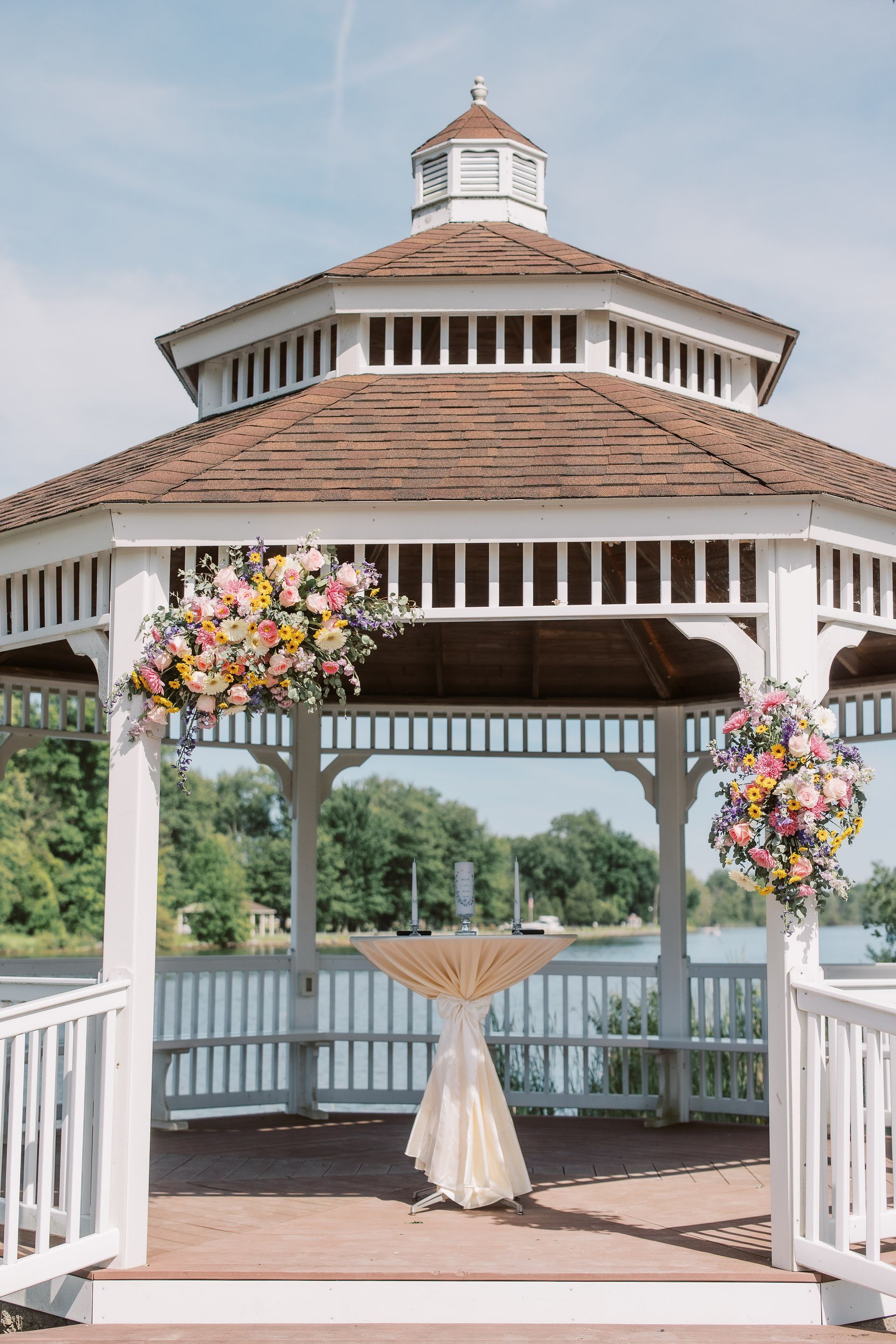 White gazebo with floral arrangements, table, and lake view.