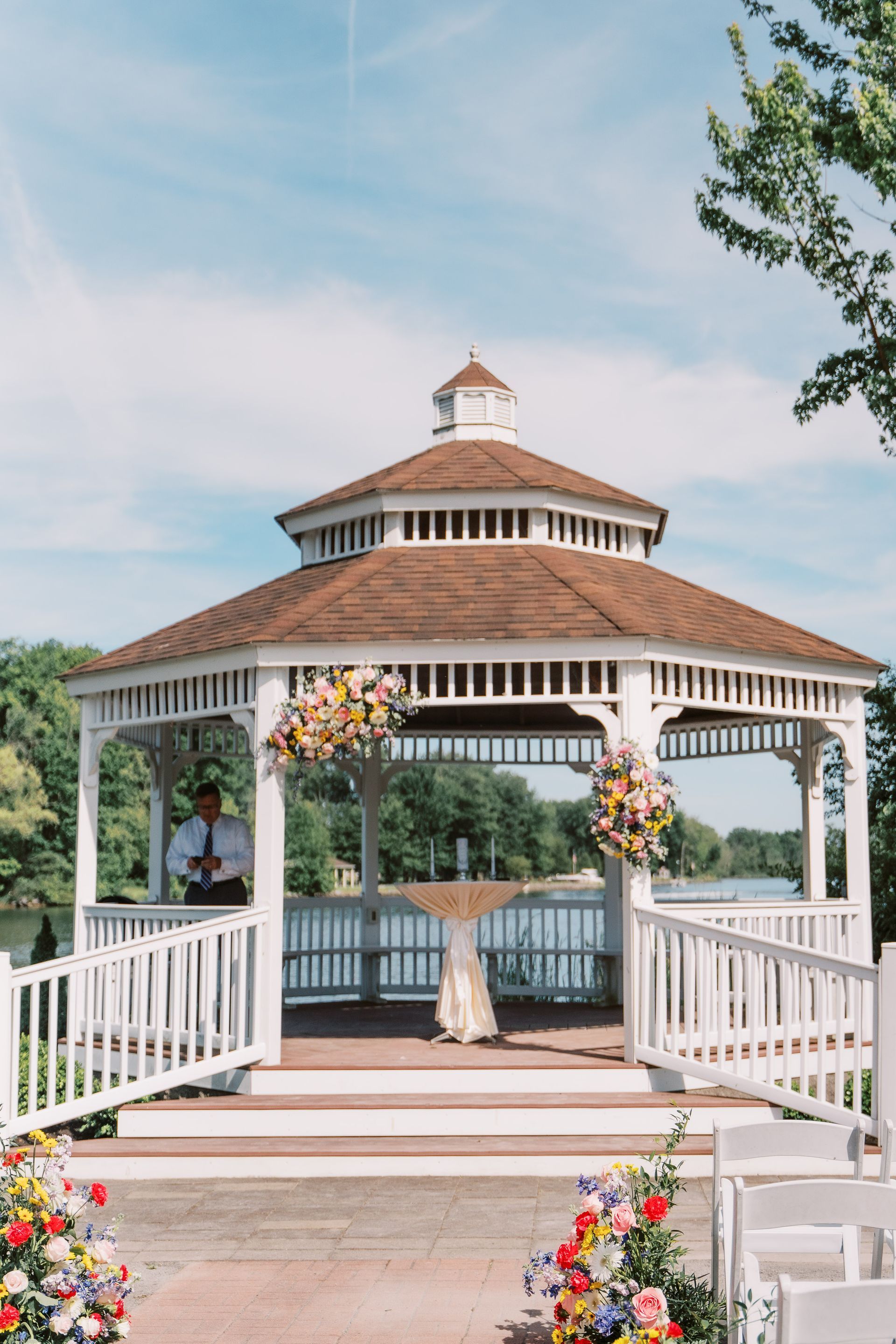 White gazebo decorated with flowers, overlooking a lake. A person stands inside the structure.