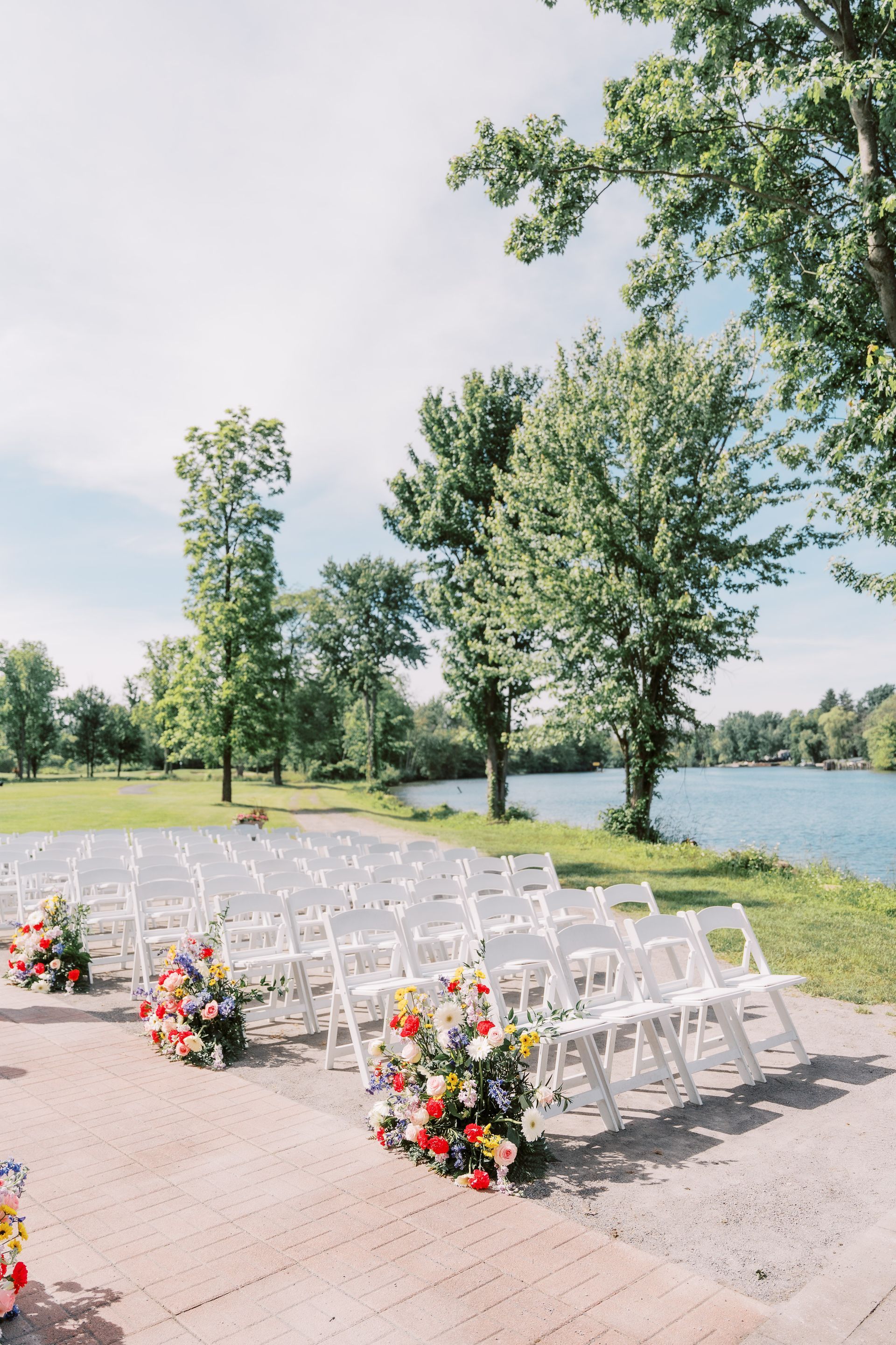White chairs arranged for a lakeside wedding ceremony; colorful floral arrangements at the end of rows; sunny day.