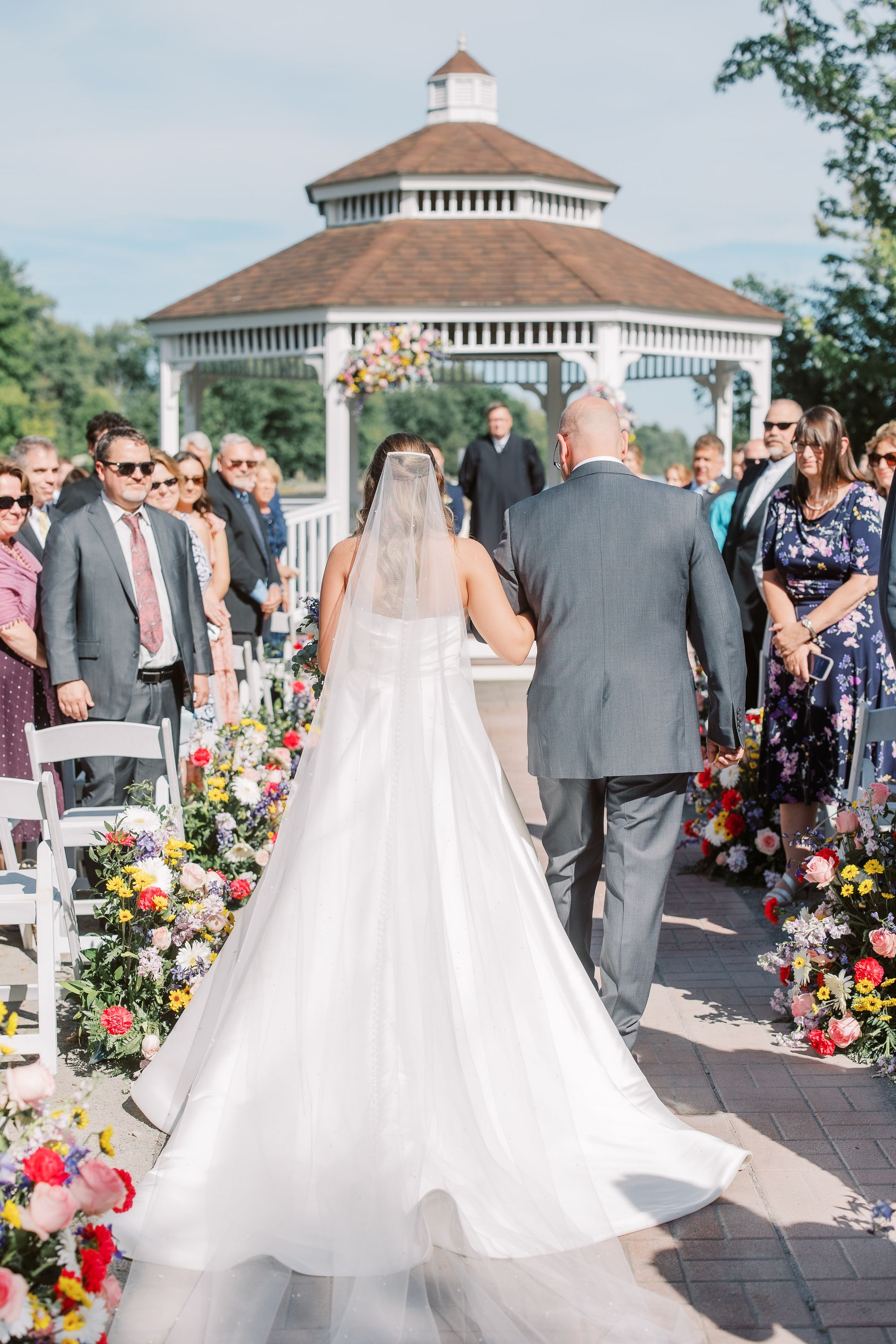 Bride walking down the aisle with her father towards a gazebo, surrounded by guests. Colorful flowers line the path.