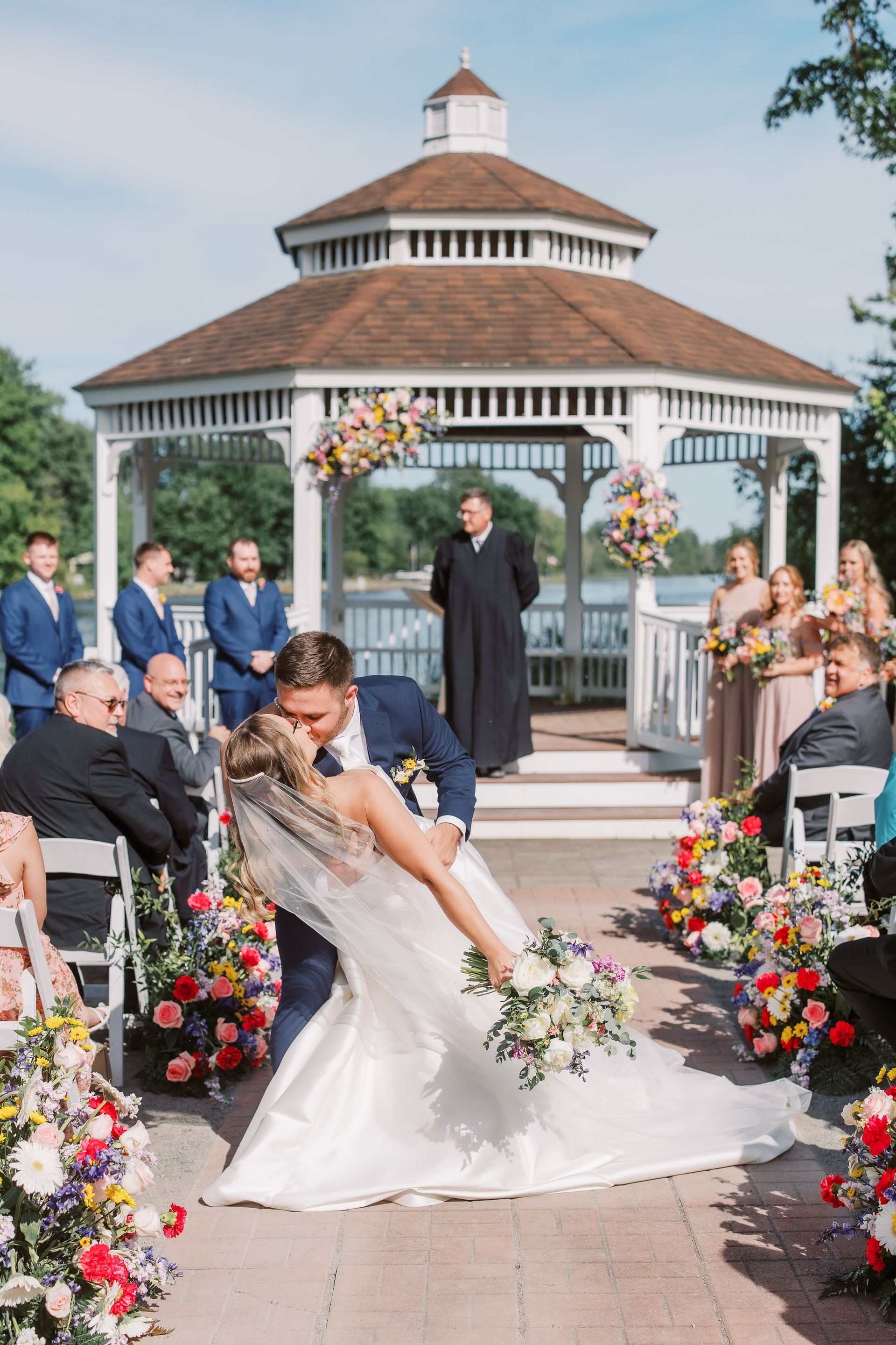 Couple kissing at outdoor wedding ceremony by a gazebo with floral decorations.