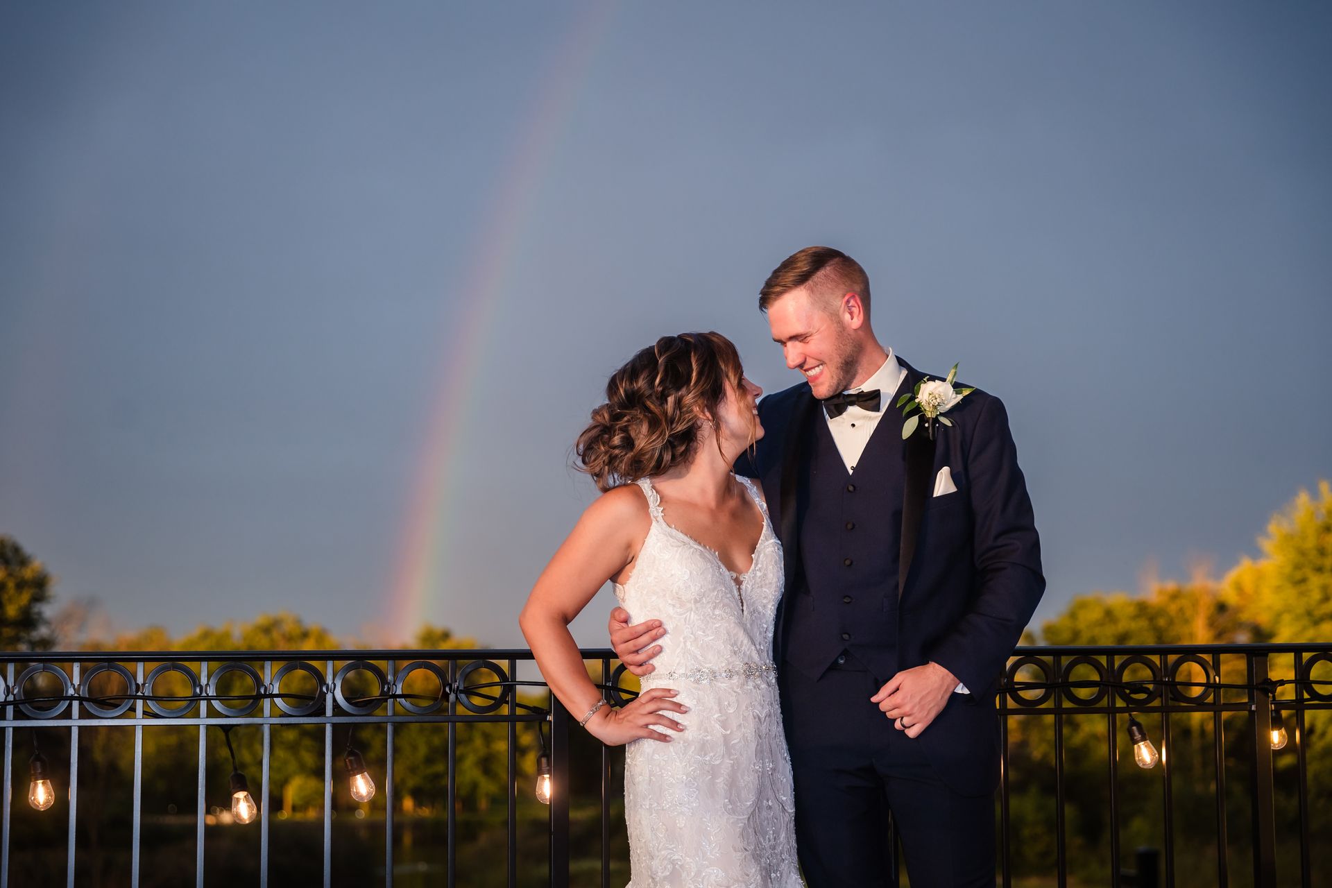 Newlyweds embrace, looking at each other, with a rainbow in the background. They are on a balcony.