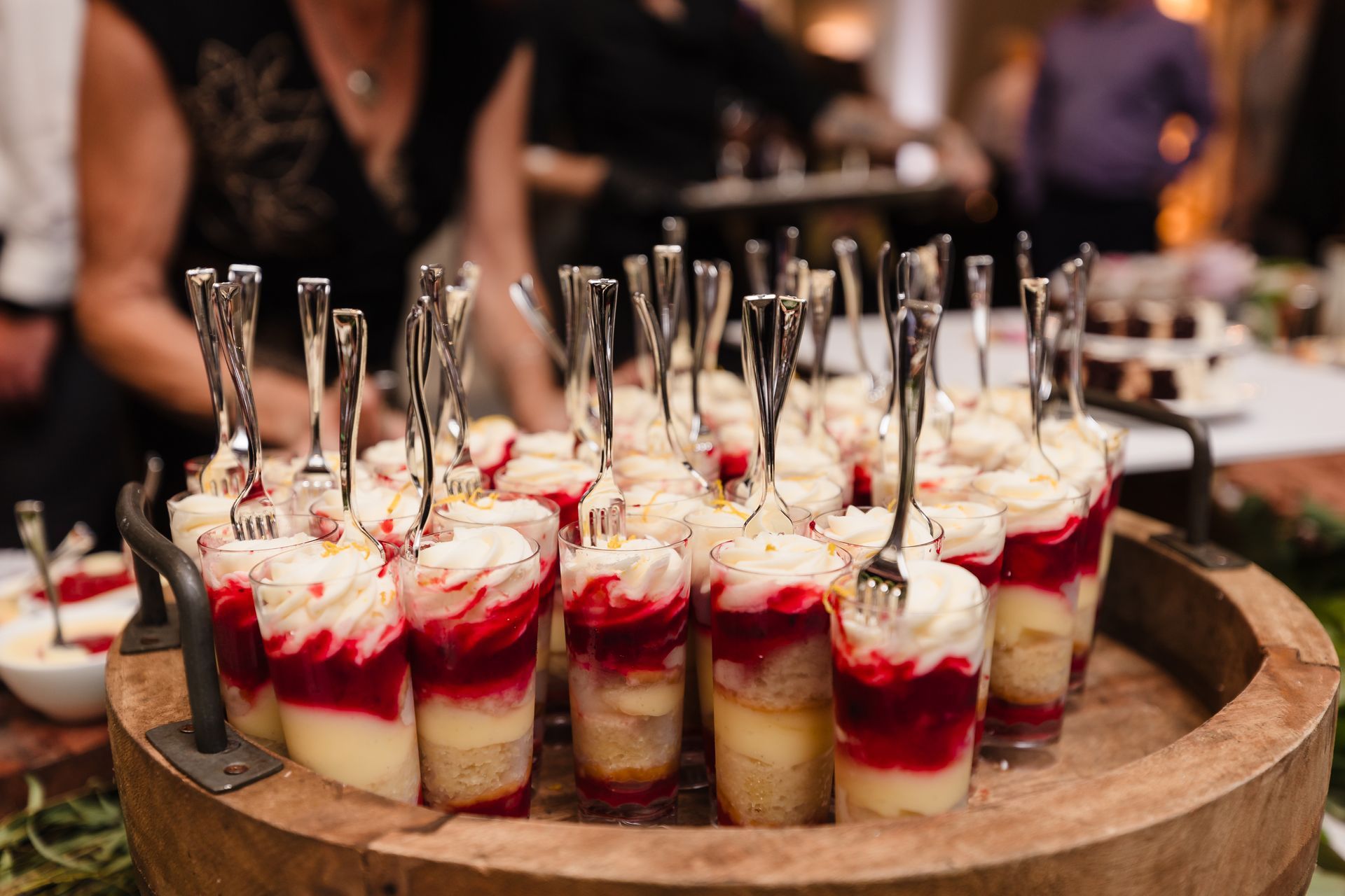 Dessert parfaits in clear cups, layered with cake, red fruit, and cream, served on a wooden platter.