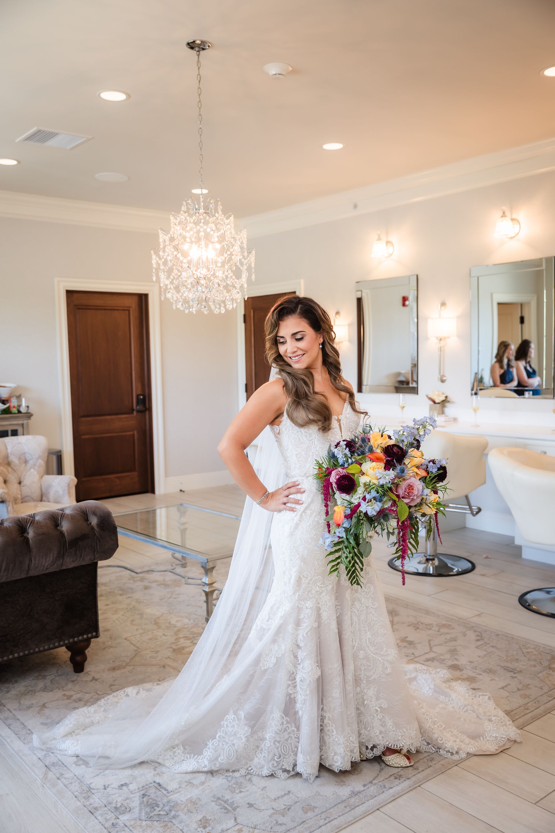 Bride in a white lace wedding dress poses holding a colorful bouquet in a well-lit room with a chandelier.