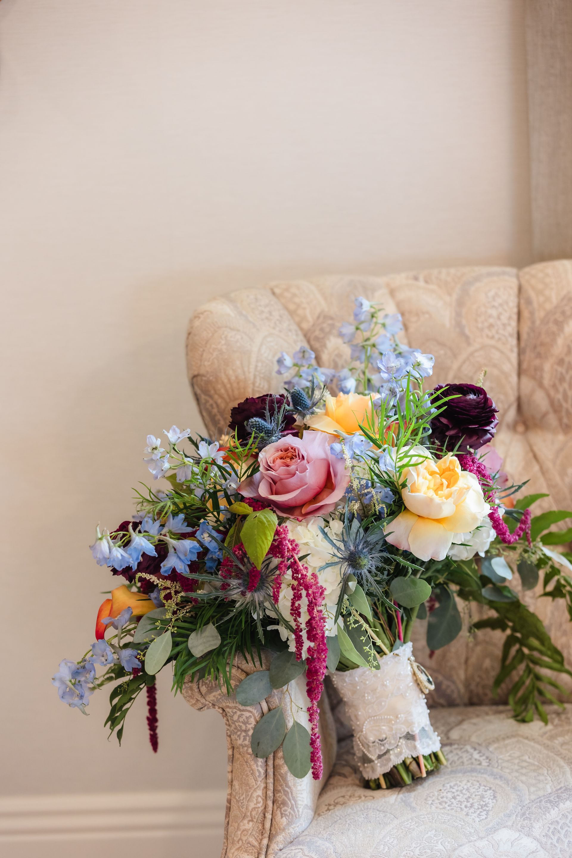 Bridal bouquet on a beige tufted chair; roses, blue delphinium, other blooms, wrapped stems.
