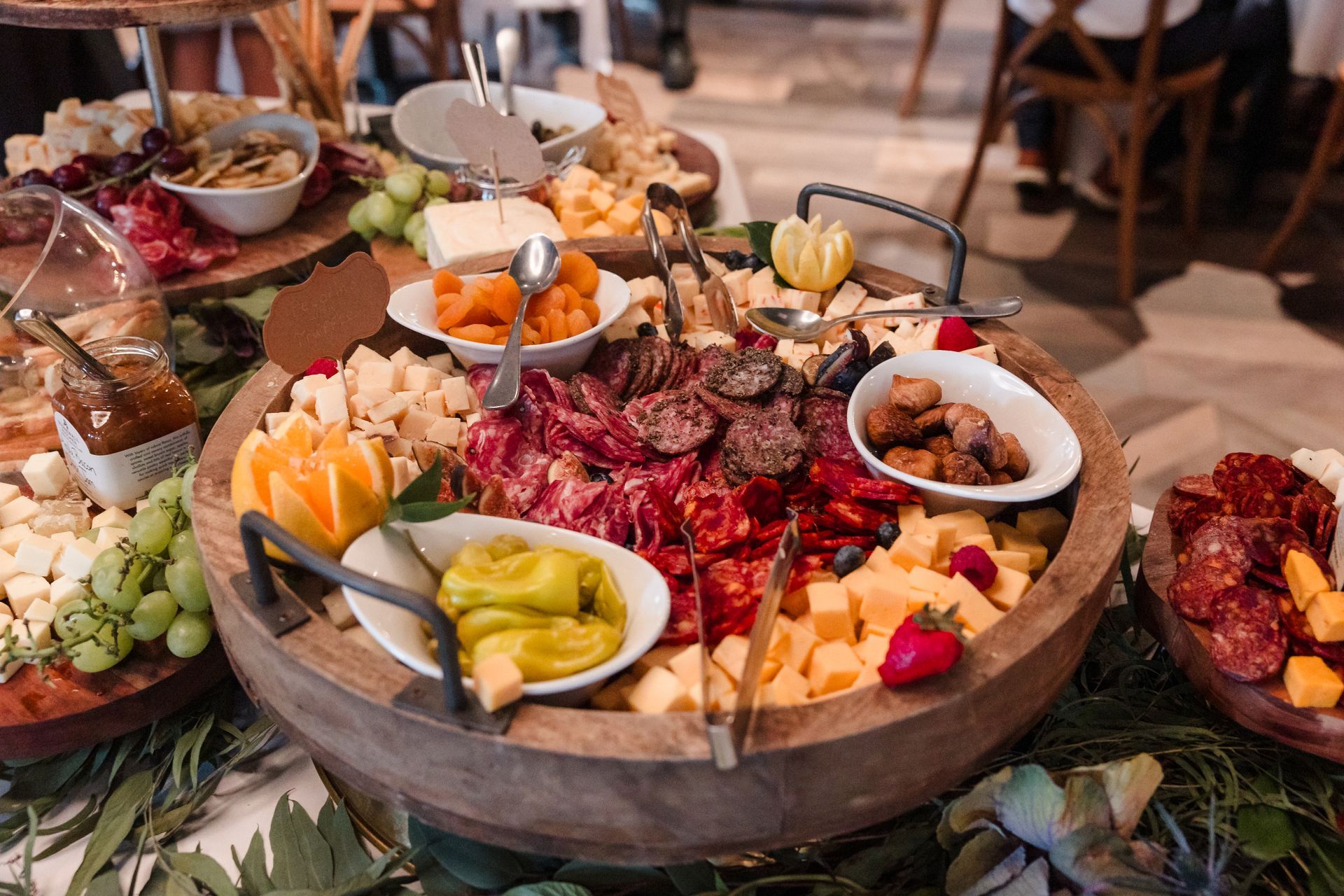 Charcuterie board with cheeses, meats, fruit, and snacks, arranged on a wooden platter with greenery.