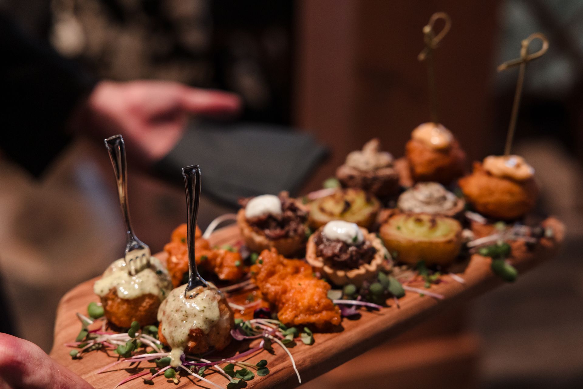 A wooden board holds an assortment of appetizers. A hand carries the board, blurred background.
