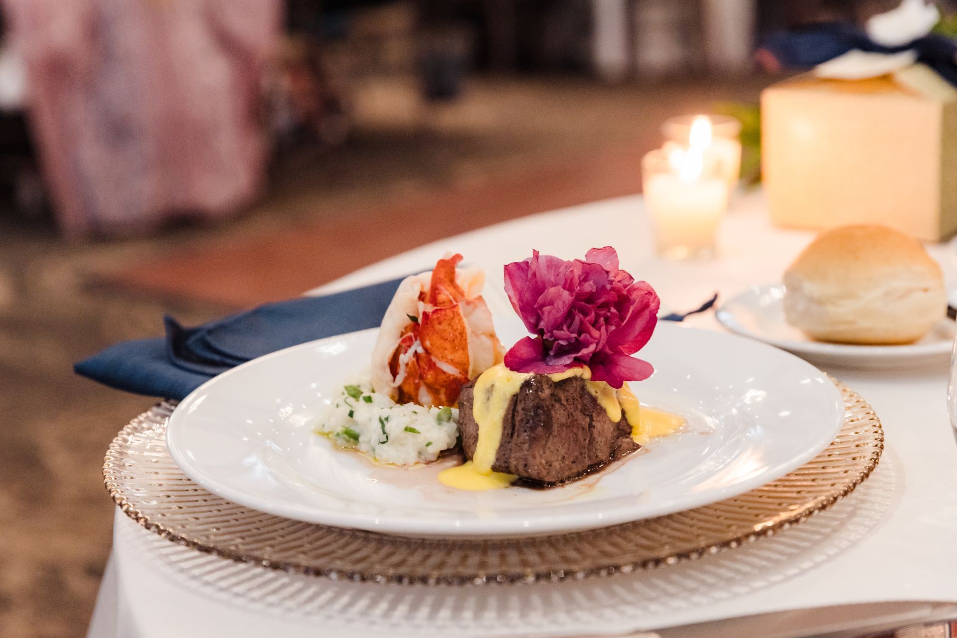 Plate of steak, lobster, and mashed potatoes, topped with a flower and sauce, next to a roll and candle.