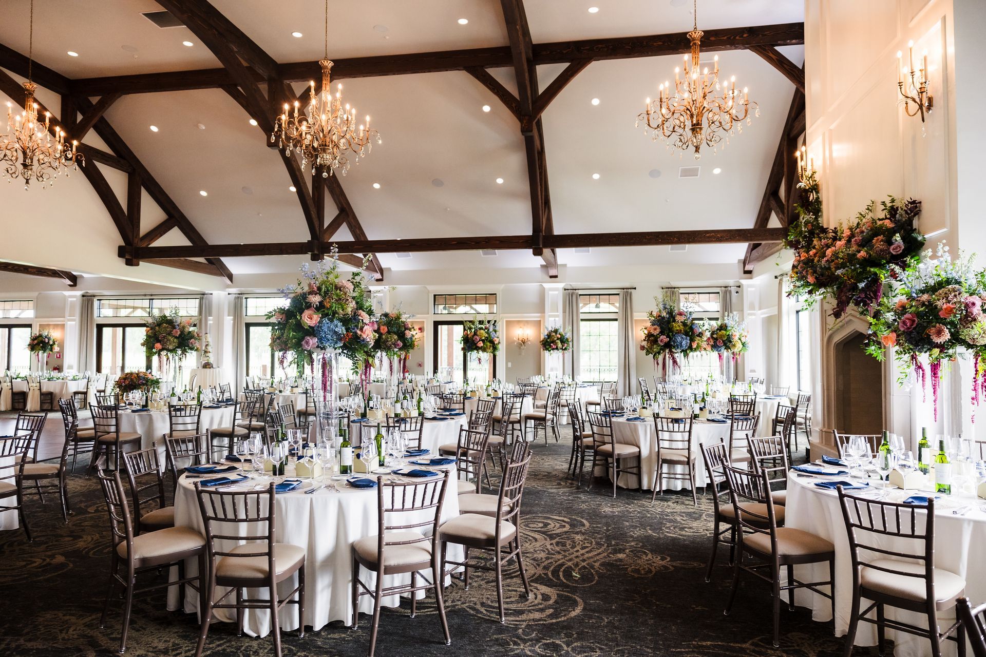 Elegant wedding reception hall with round tables, floral centerpieces, chandeliers, and exposed wooden beams.
