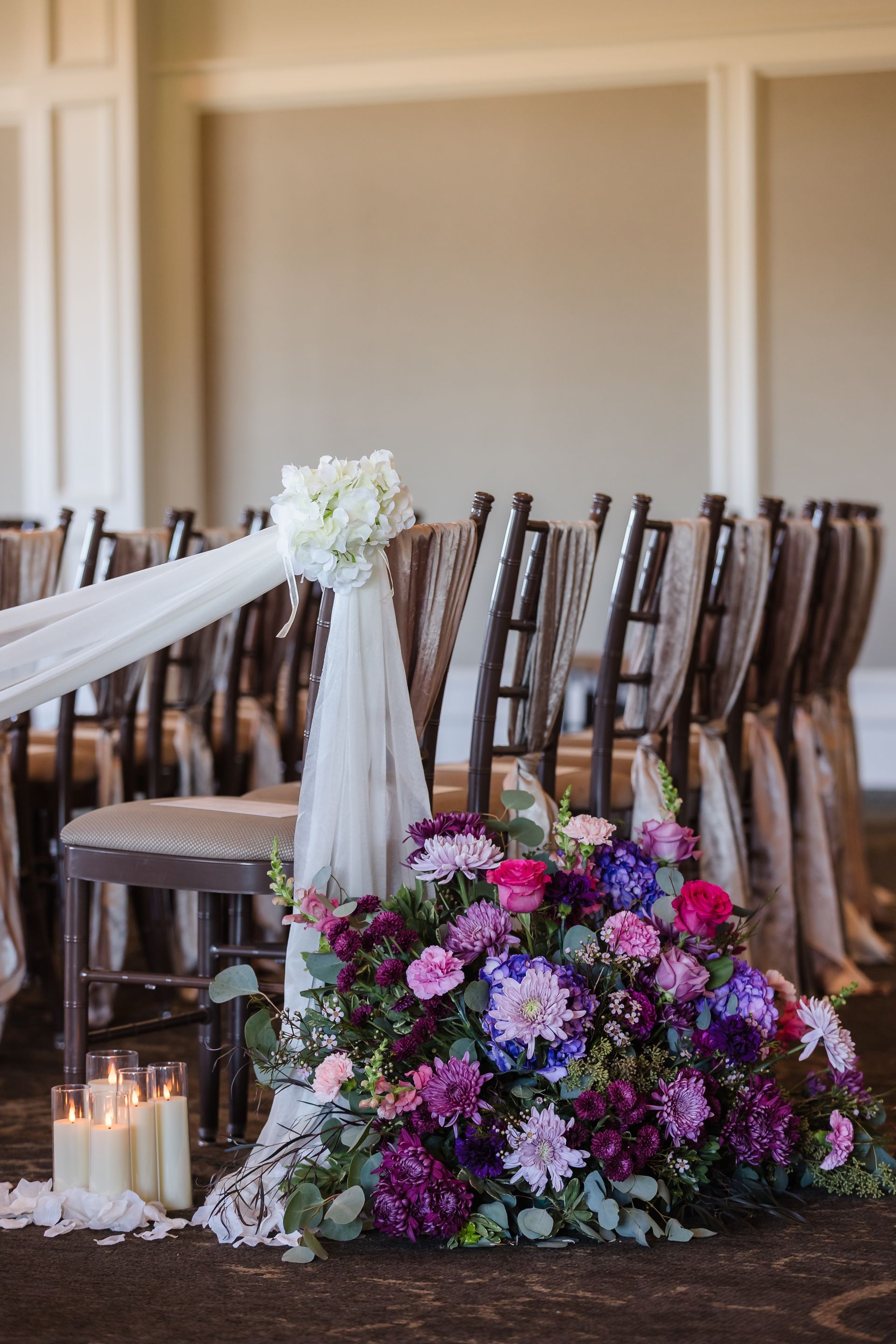 Wedding aisle decor: floral arrangement with purple and pink flowers, draped chair with white flowers and sheer fabric, candles.