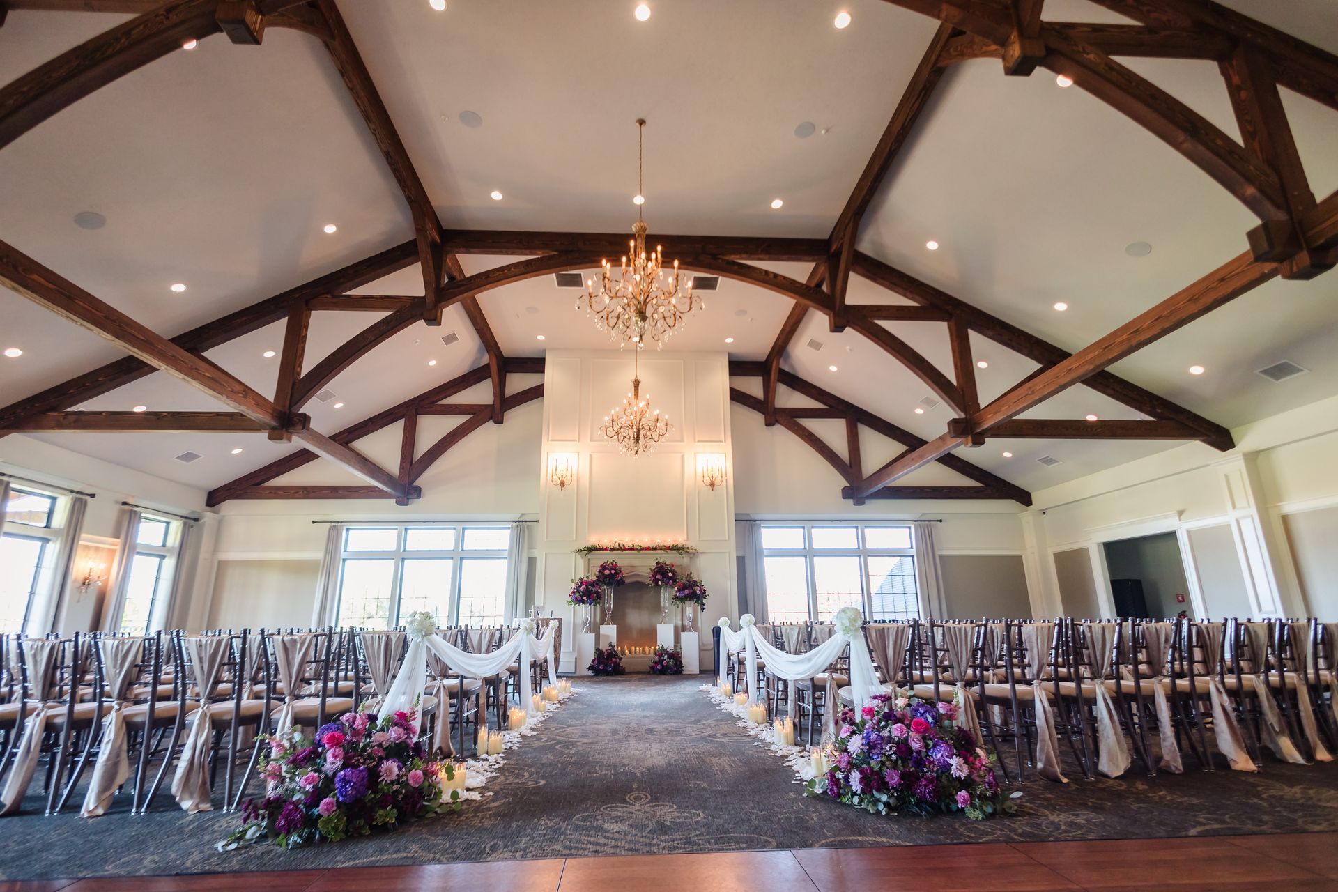 Wedding ceremony setup in a ballroom; chairs face an altar, floral arrangements, chandeliers, and high wood beam ceiling.