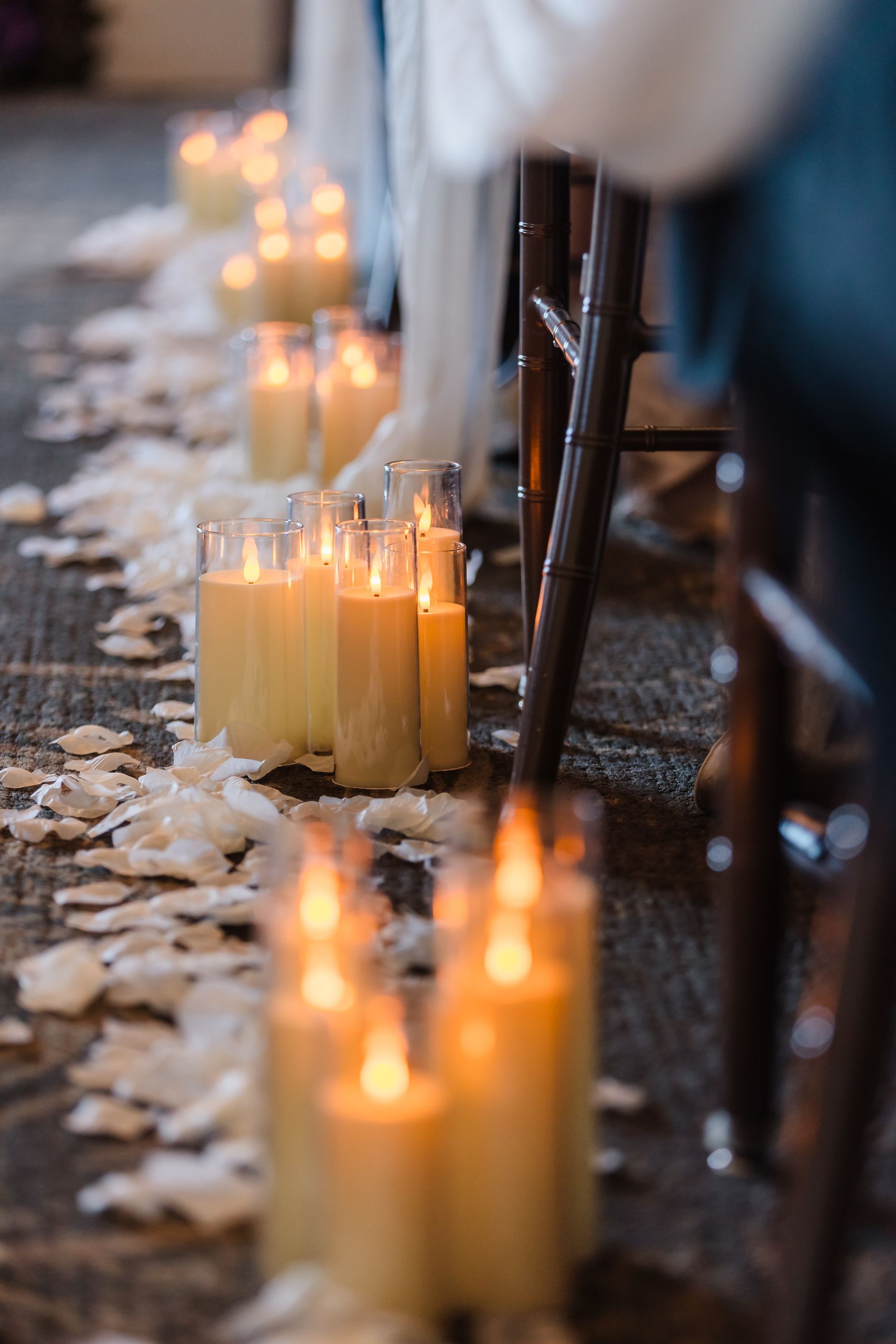 Candlelit aisle with scattered white petals, viewed from the side, near wooden chairs and draped fabric.