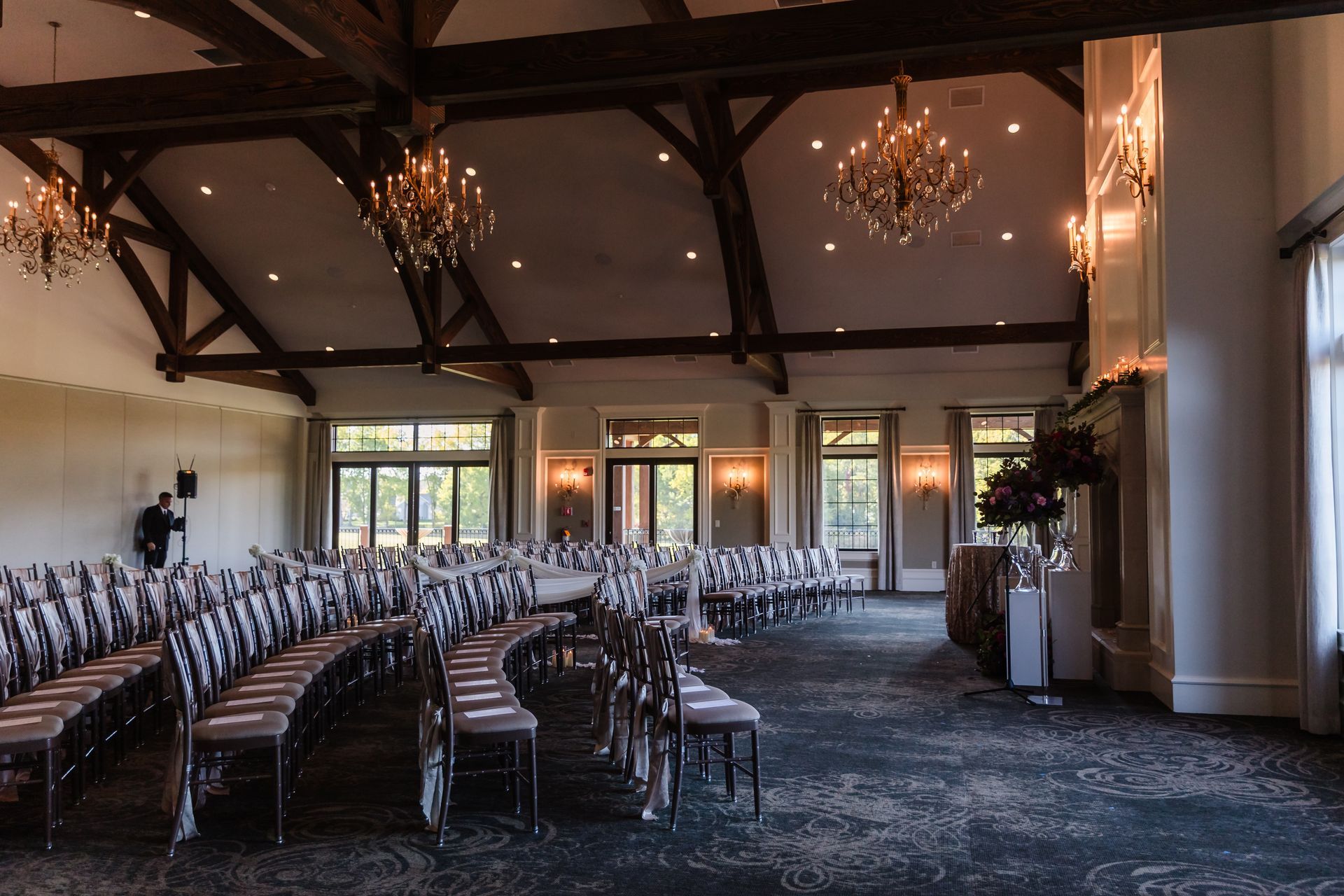 Empty room set up for an event, featuring curved rows of chairs, chandeliers, and large windows.
