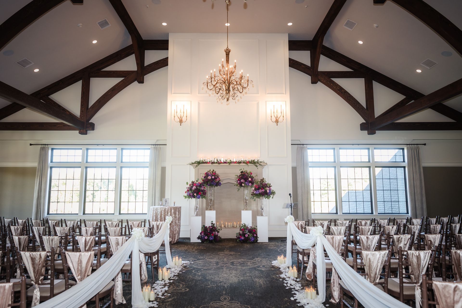 Indoor wedding ceremony setup, altar with floral arrangements, rows of chairs, sheer fabric aisle runner, and overhead chandelier.